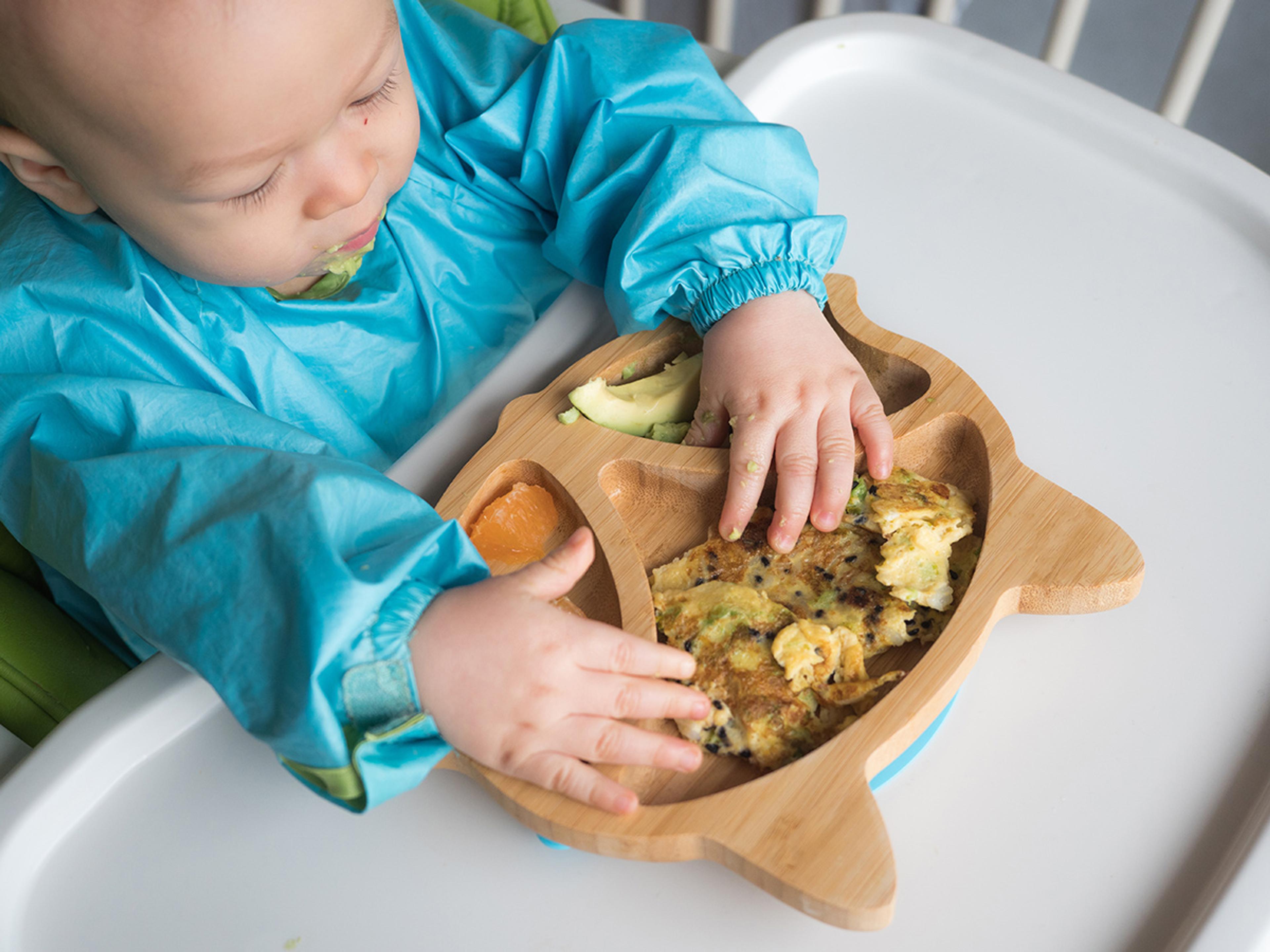 Asian baby eats a plate of food, including avocados and scrambled eggs.