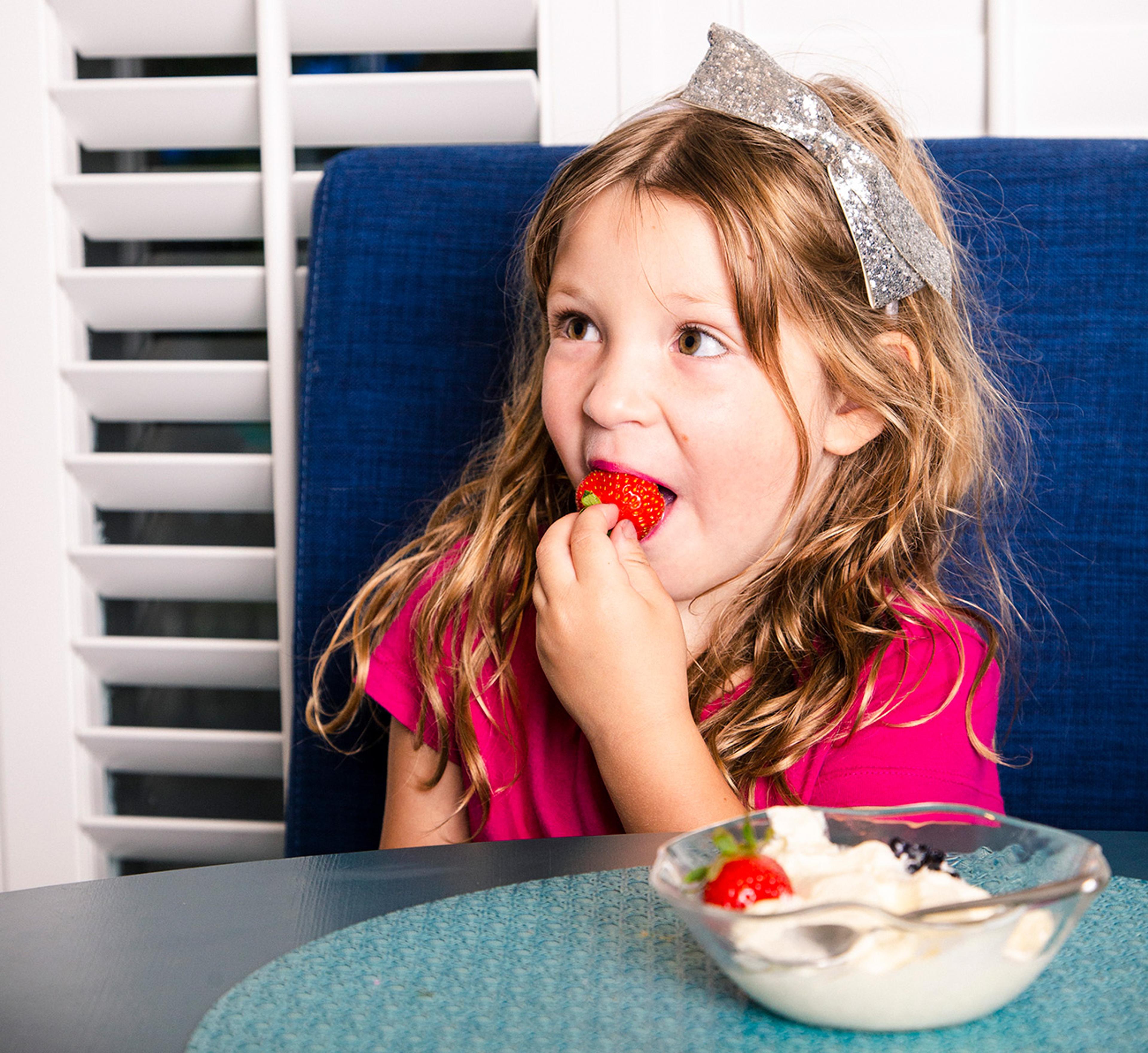 A young girl wearing a pink shirt eats a strawberry with whipped cream.