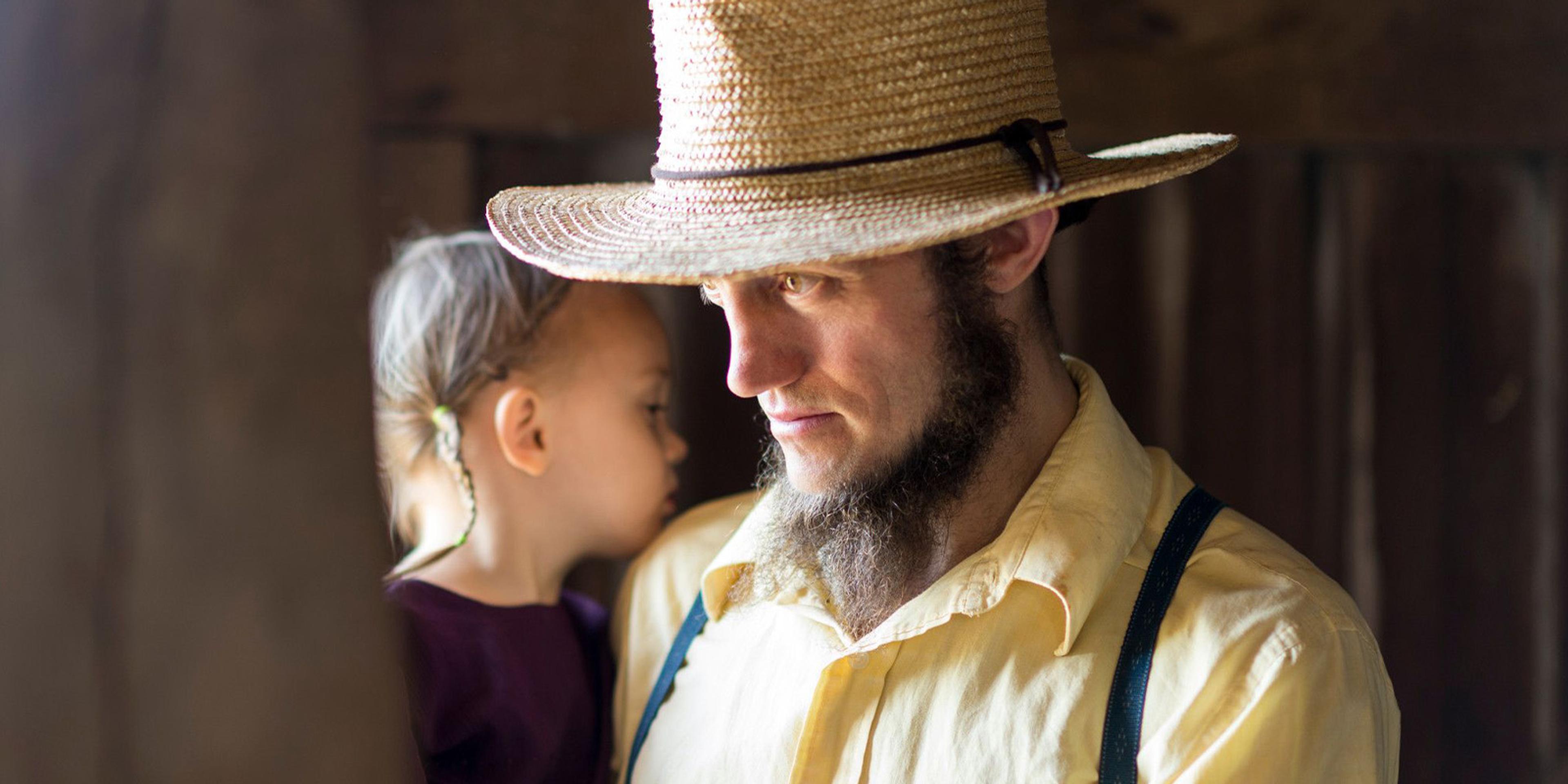 A farmer wearing a straw hat and suspenders holds a girl with braided hair.