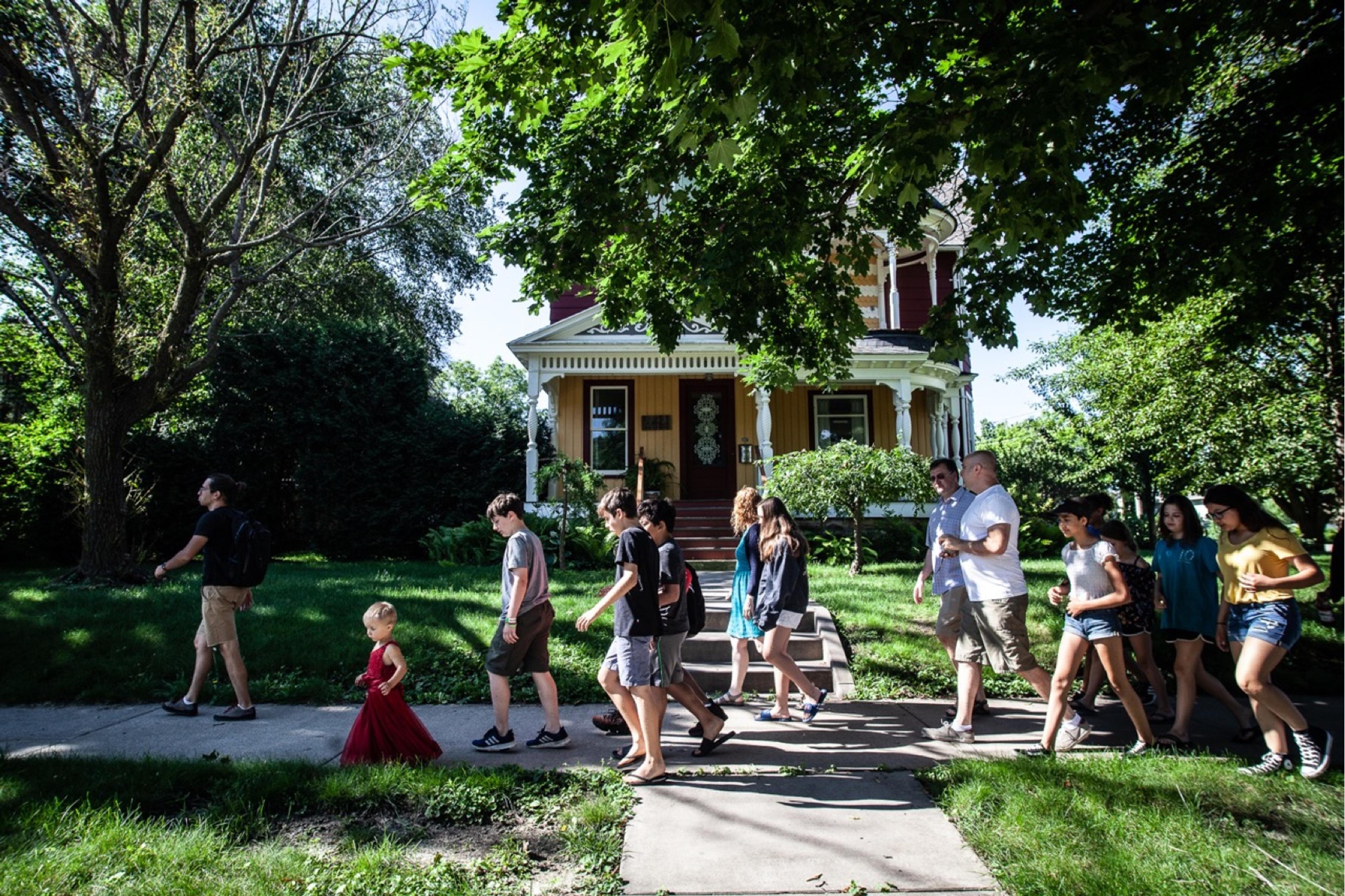 A group of young people walk along a sidewalk in front of a large yellow house.