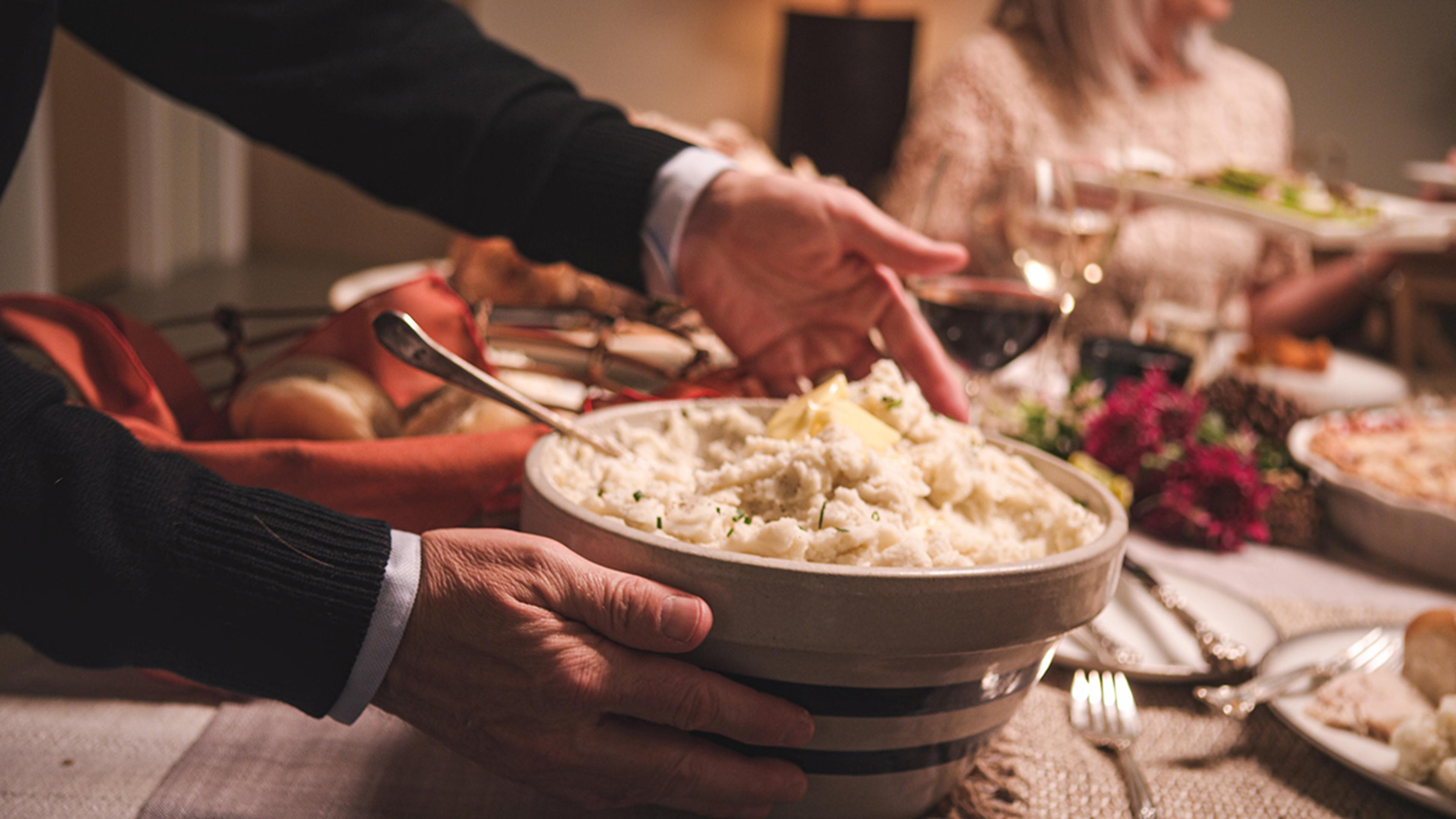 A man places a large bowl of mashed potatoes topped with butter on a Thanksgiving table surrounded by people.