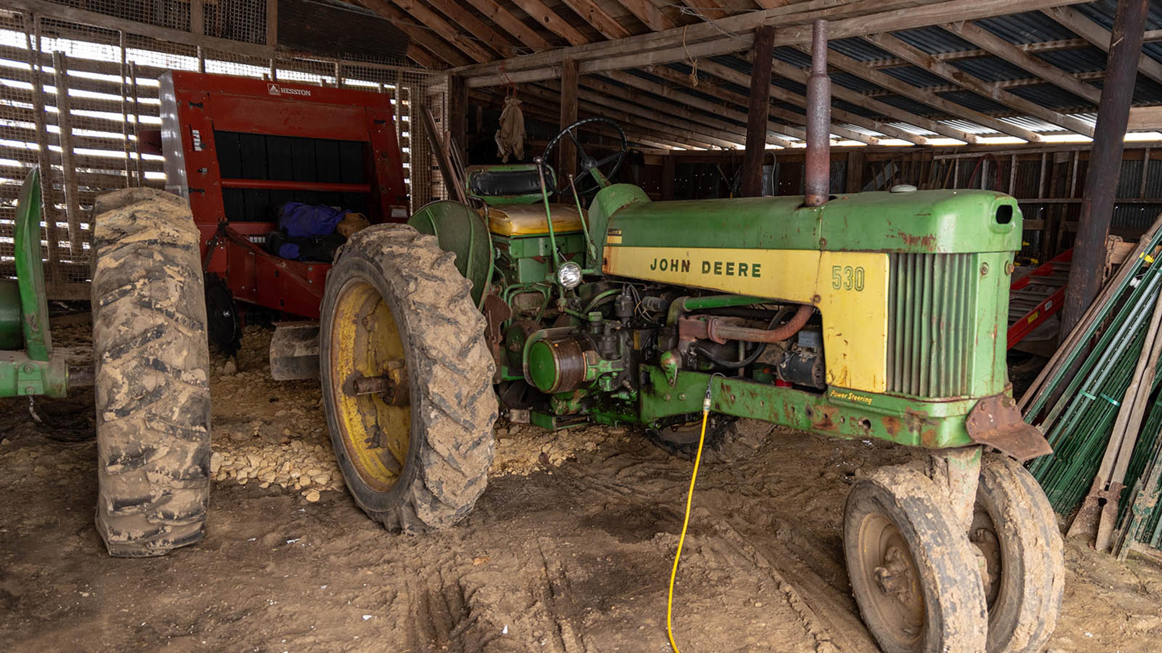 Old green John Deere diesel tractor in the barn with an electrical cord coming out of the engine.