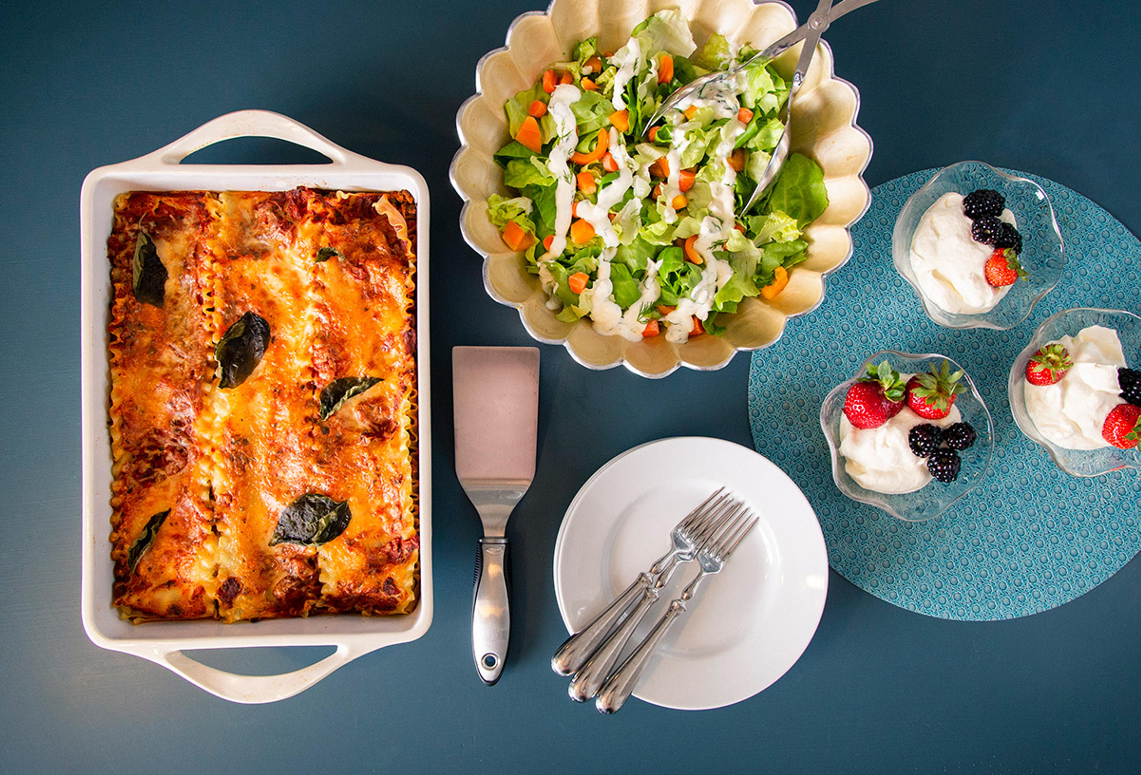 A pan of lasagna, salad, and dishes of fruit with whipped cream arranged around plates and forks on a blue table.
