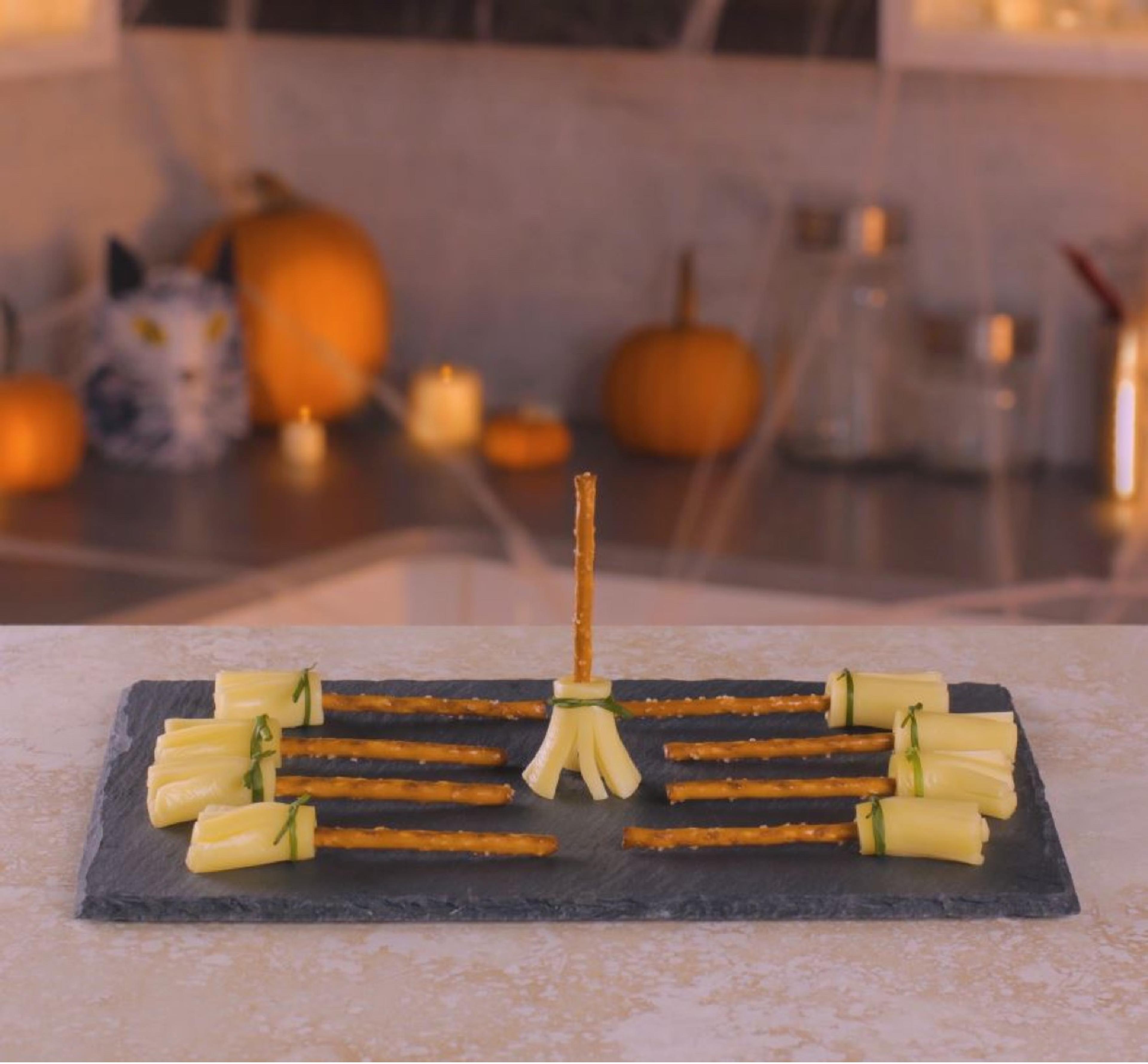 Nine fully assembled string cheese pretzel broomsticks displayed on a slate tray with fall decorations in the background.