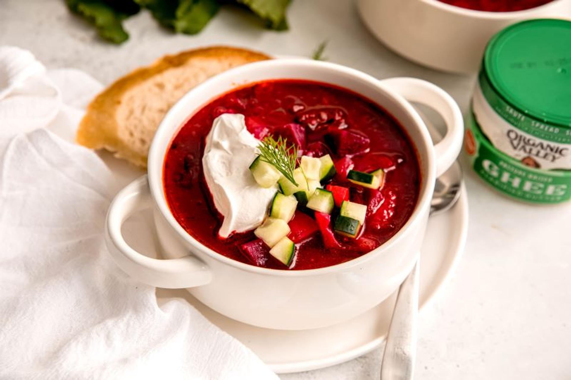 A beautiful bowl of borscht with cucumbers and sour cream on a table next to bread and a jar of organic ghee.