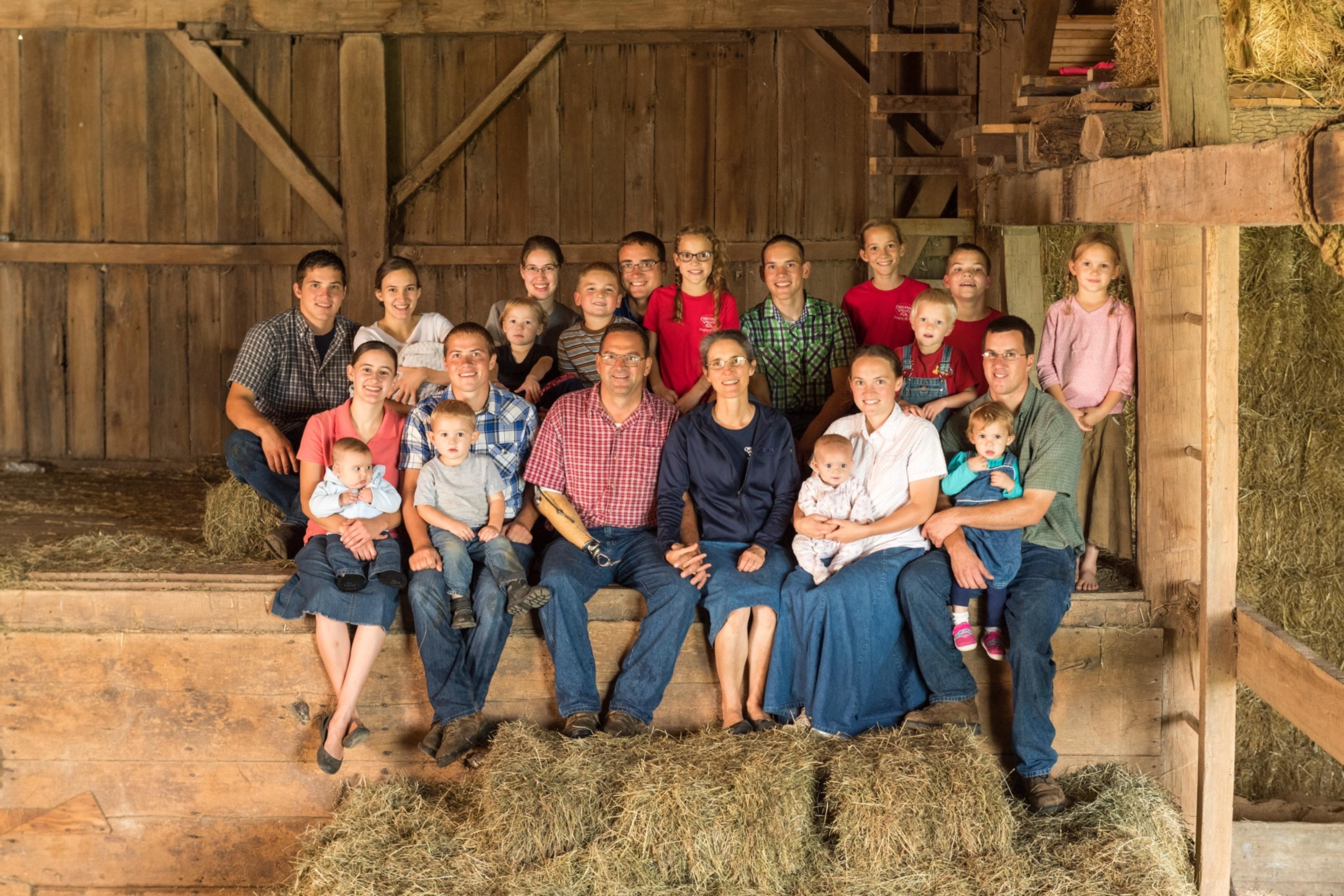 A portrait of a farm family made up of 23 adults, children and infants seated in their barn surrounded by hay bales.