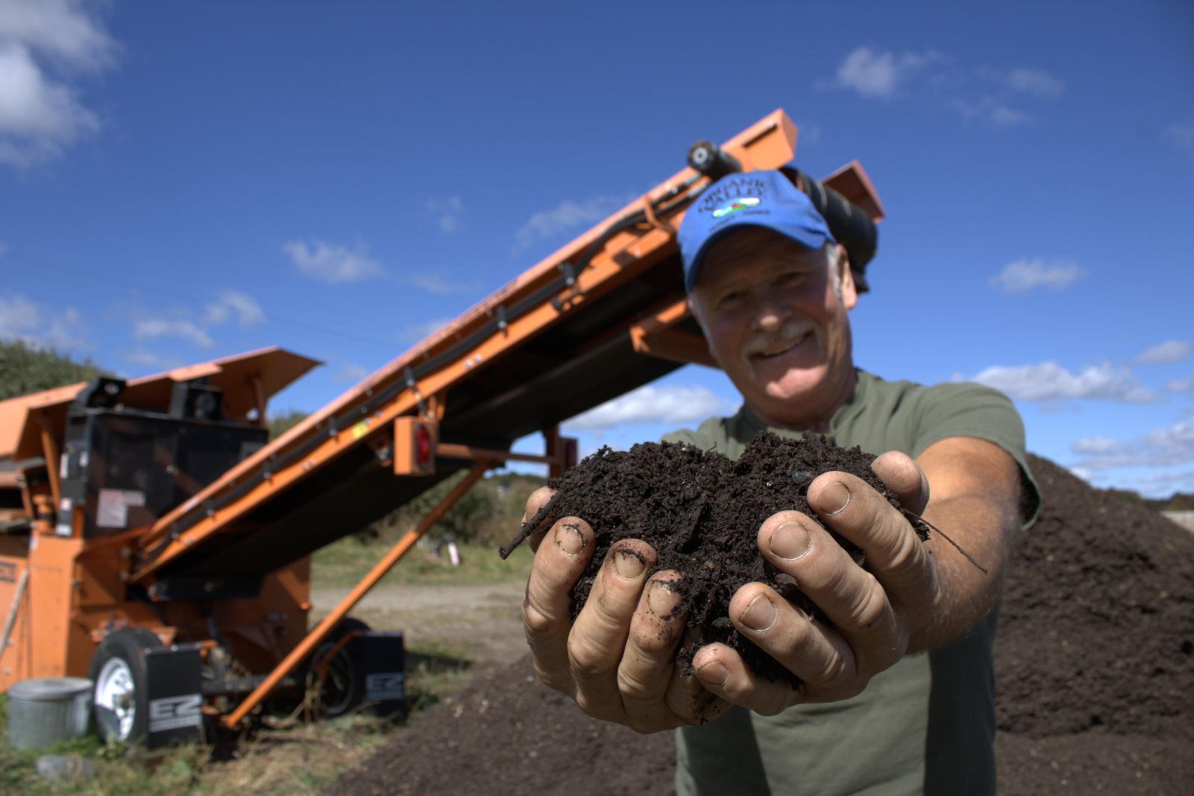 Eric Paris, Vermont, holds two handfuls of compost while standing in front of a screening implement and compost pile.