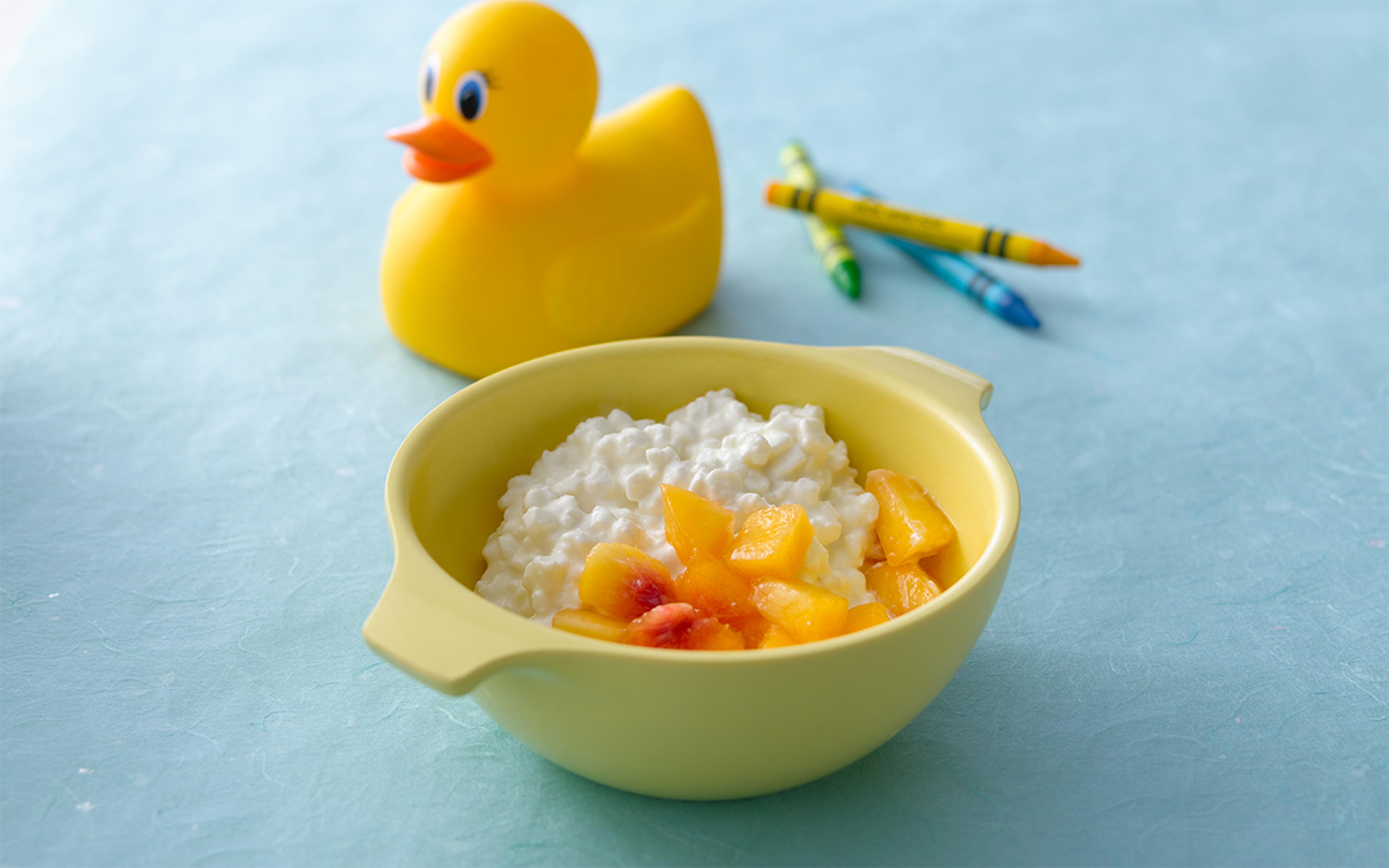 A cup of cottage cheese and cut peaches with a rubber ducky and some crayons in the background.