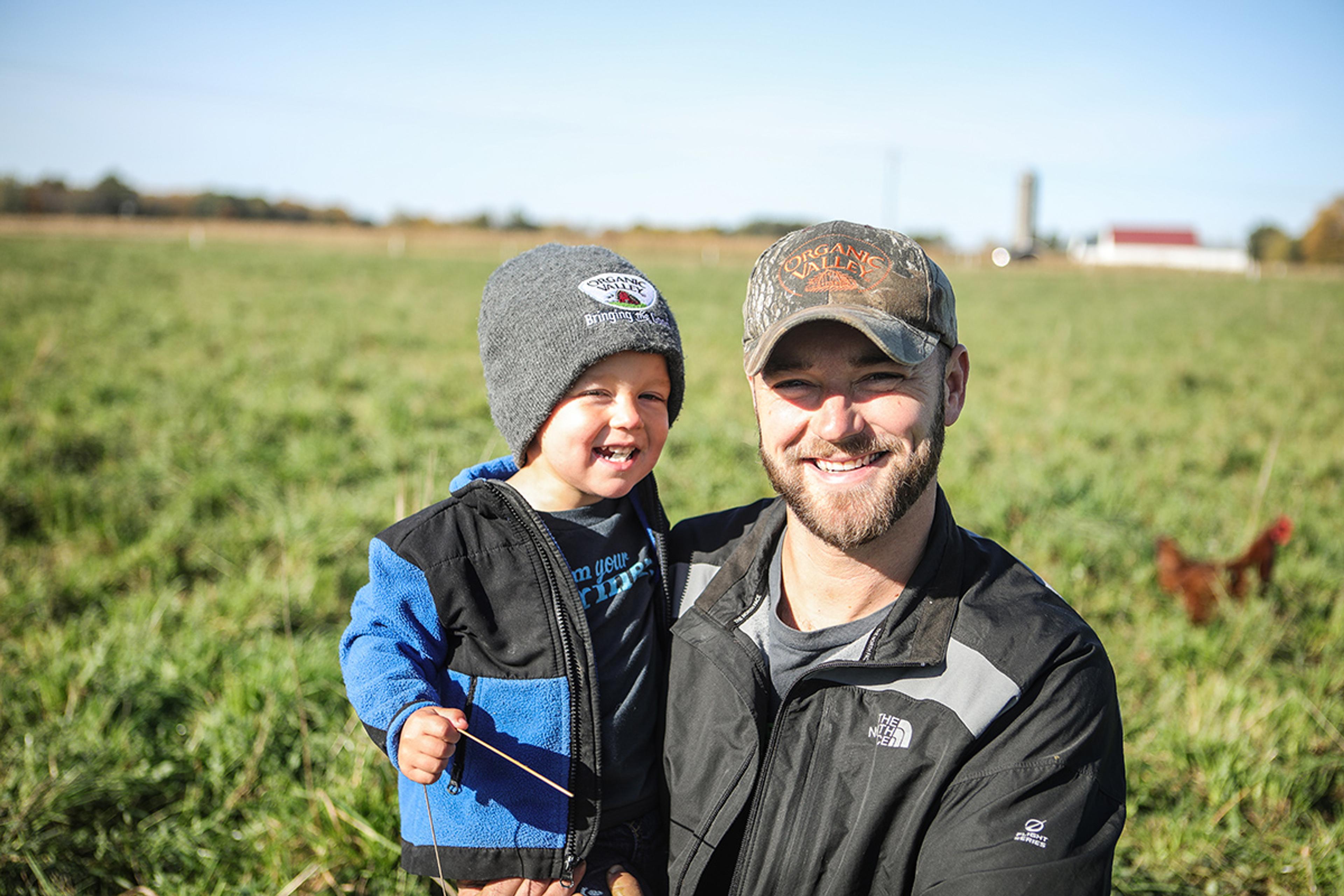 Organic Valley farmer, Jordan Settlage smiles holding his son in the pasture of his farm.