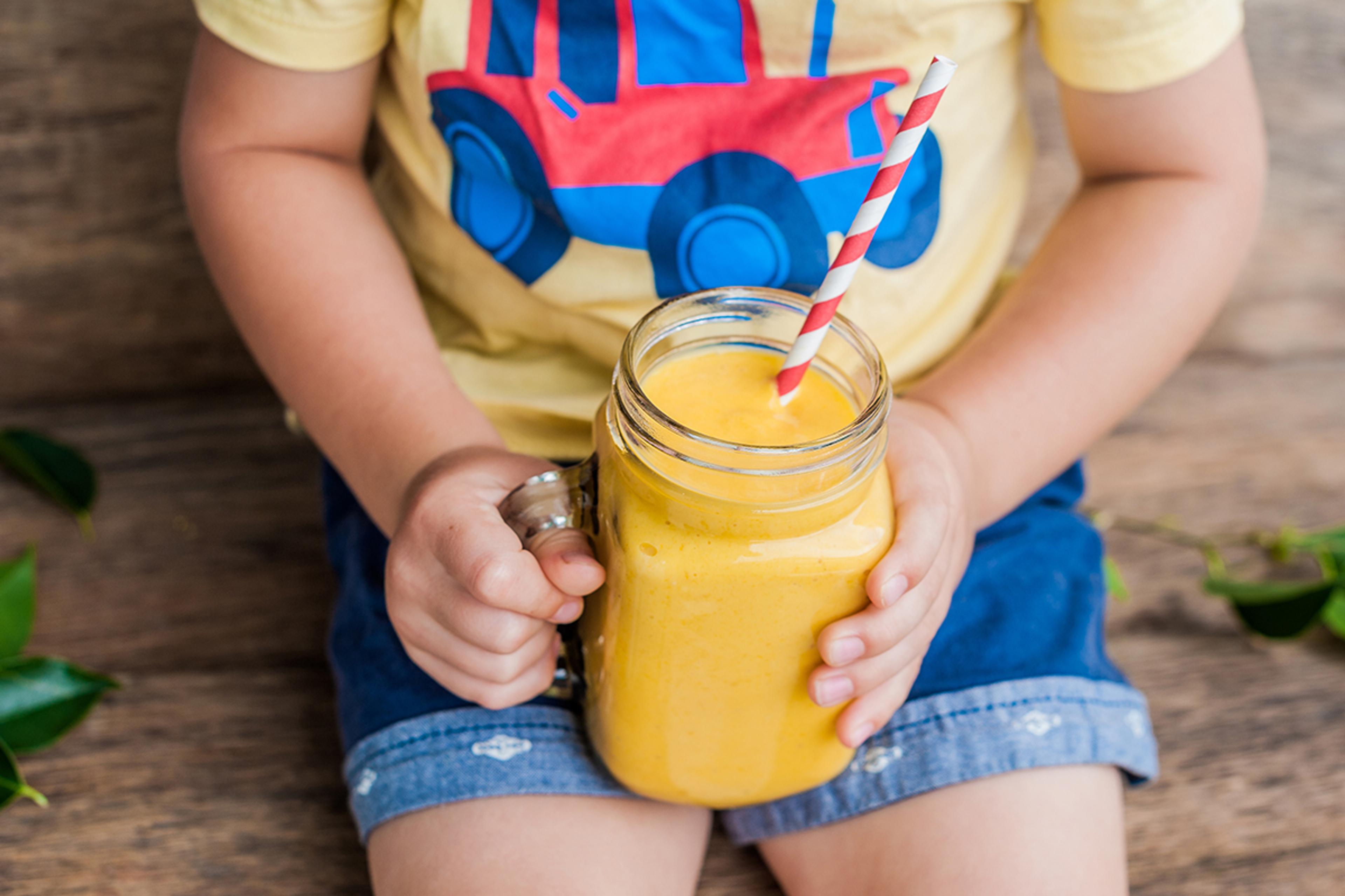 A child holds a cup containing a bright yellow smoothie with a red and white striped straw.