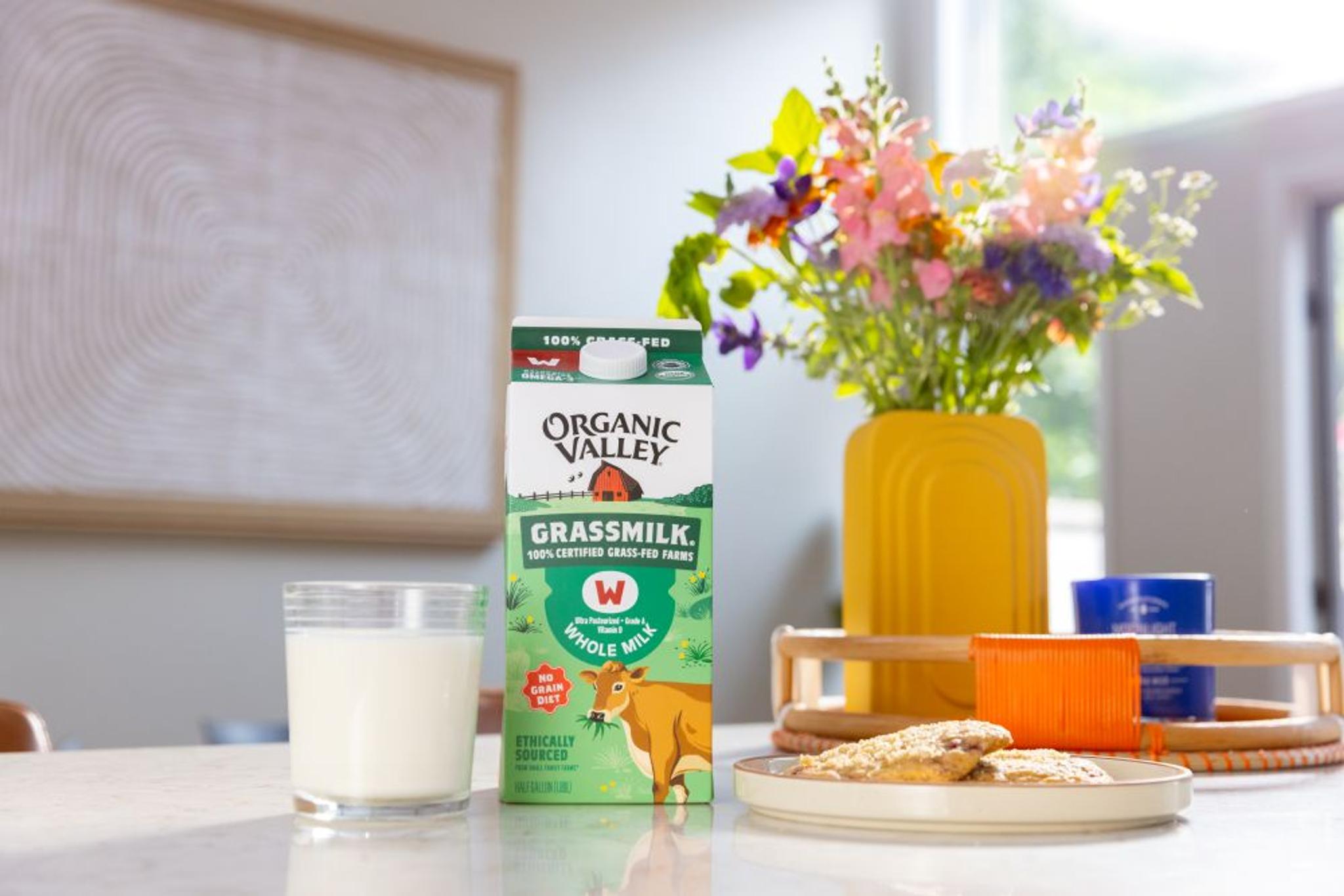 Glass of milk next to a carton of Grassmilk® Milk, with a plate of cookies and vase of flowers nearby.
