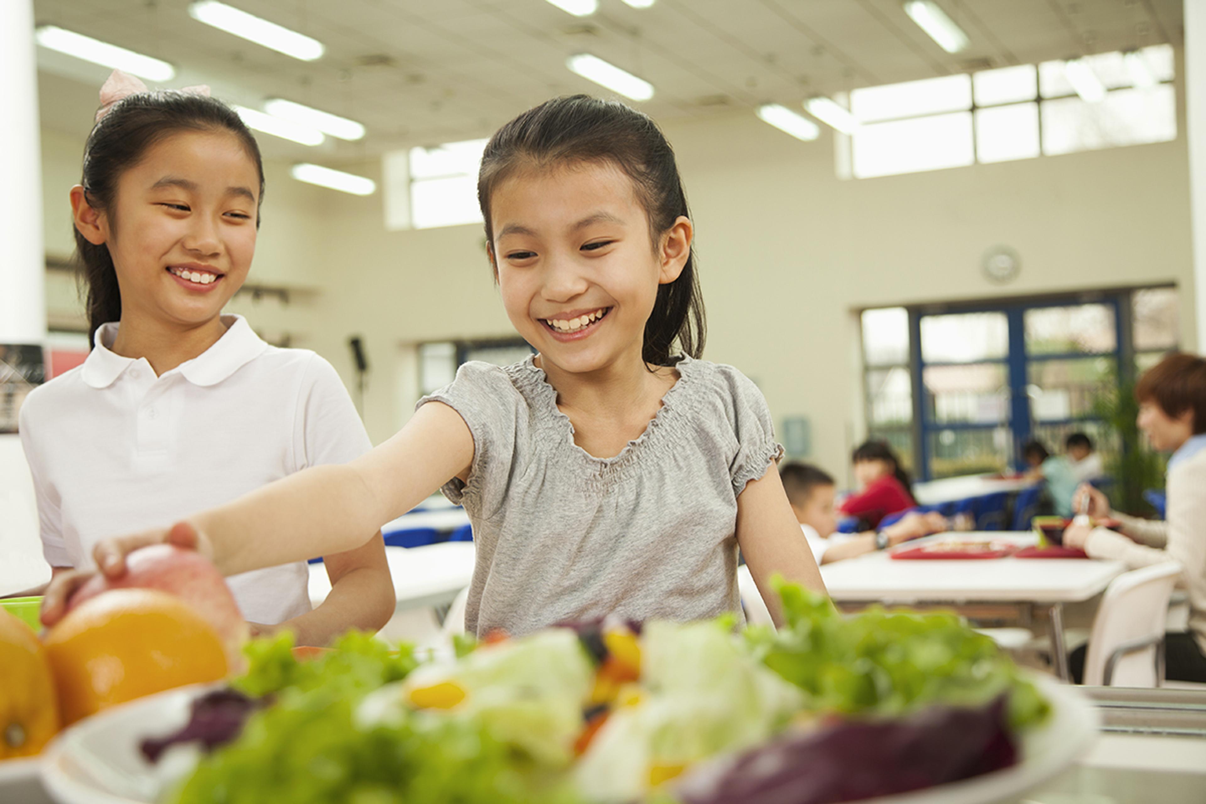 Two young girls smile while taking fresh foods from their school lunch salad bar.