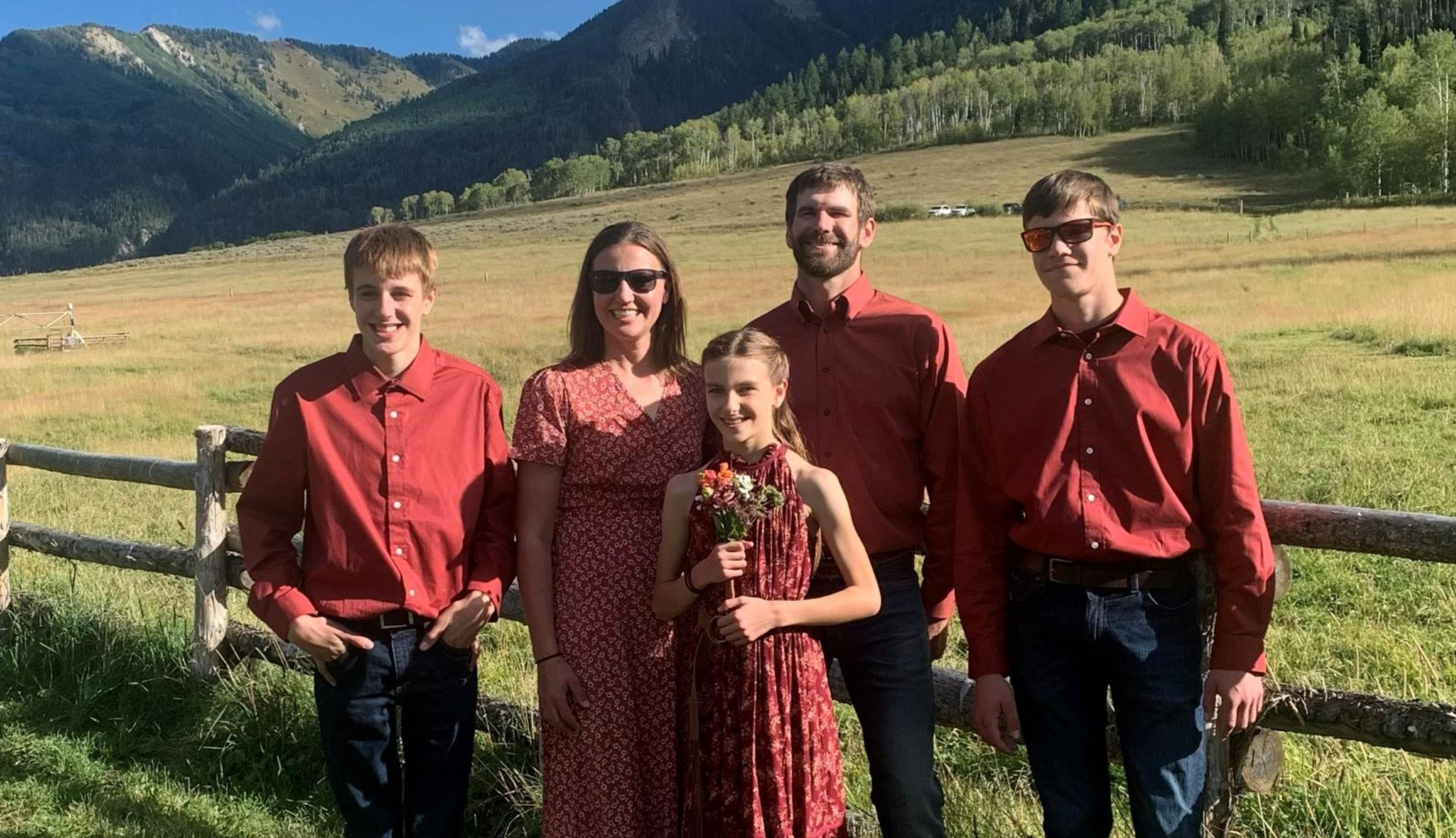 The five members of the Zweber family stand in front of a fence with a grassy field and hills in the background.