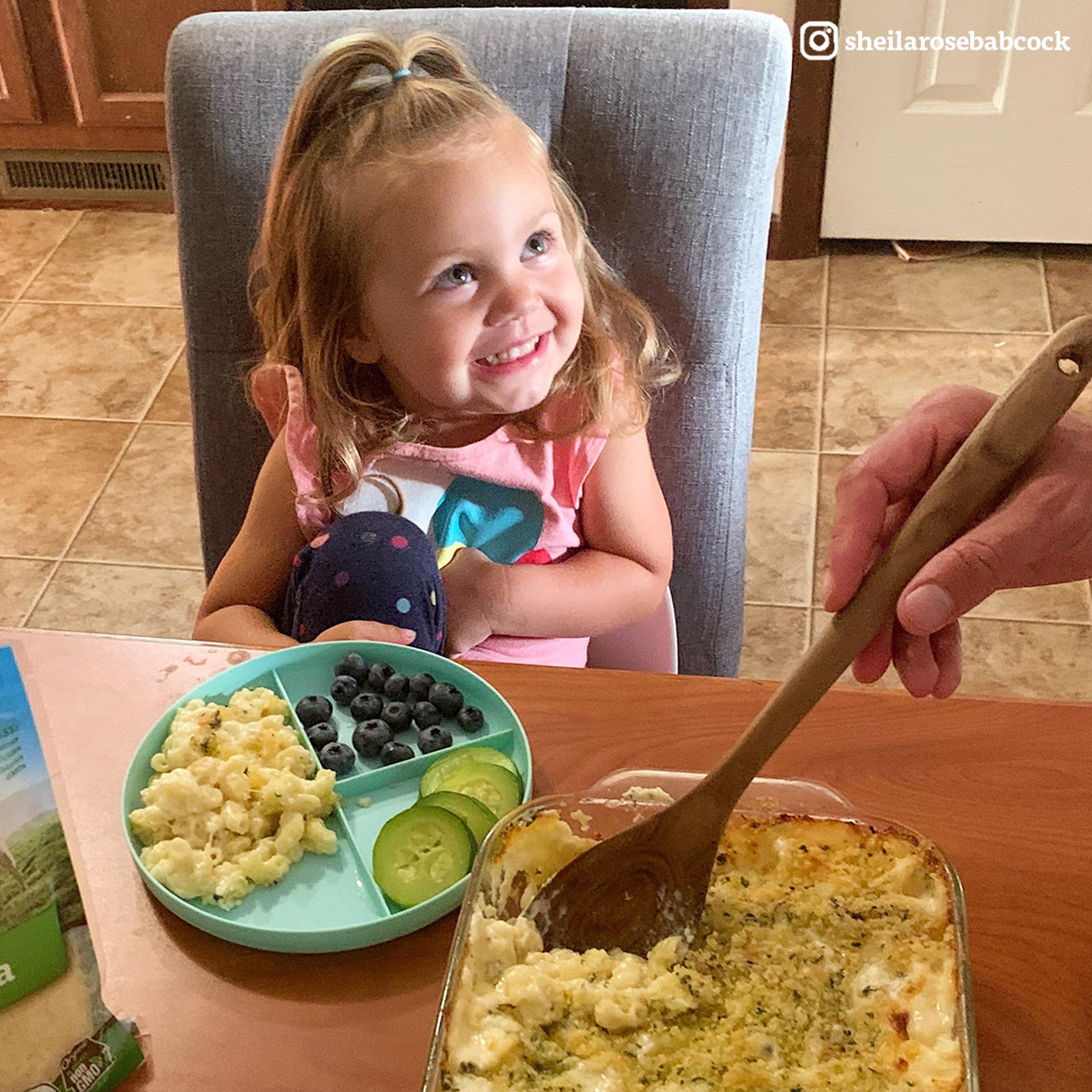 A girl sits at the dinner table with a plate of organic veggies and cheesy noodle casserole.