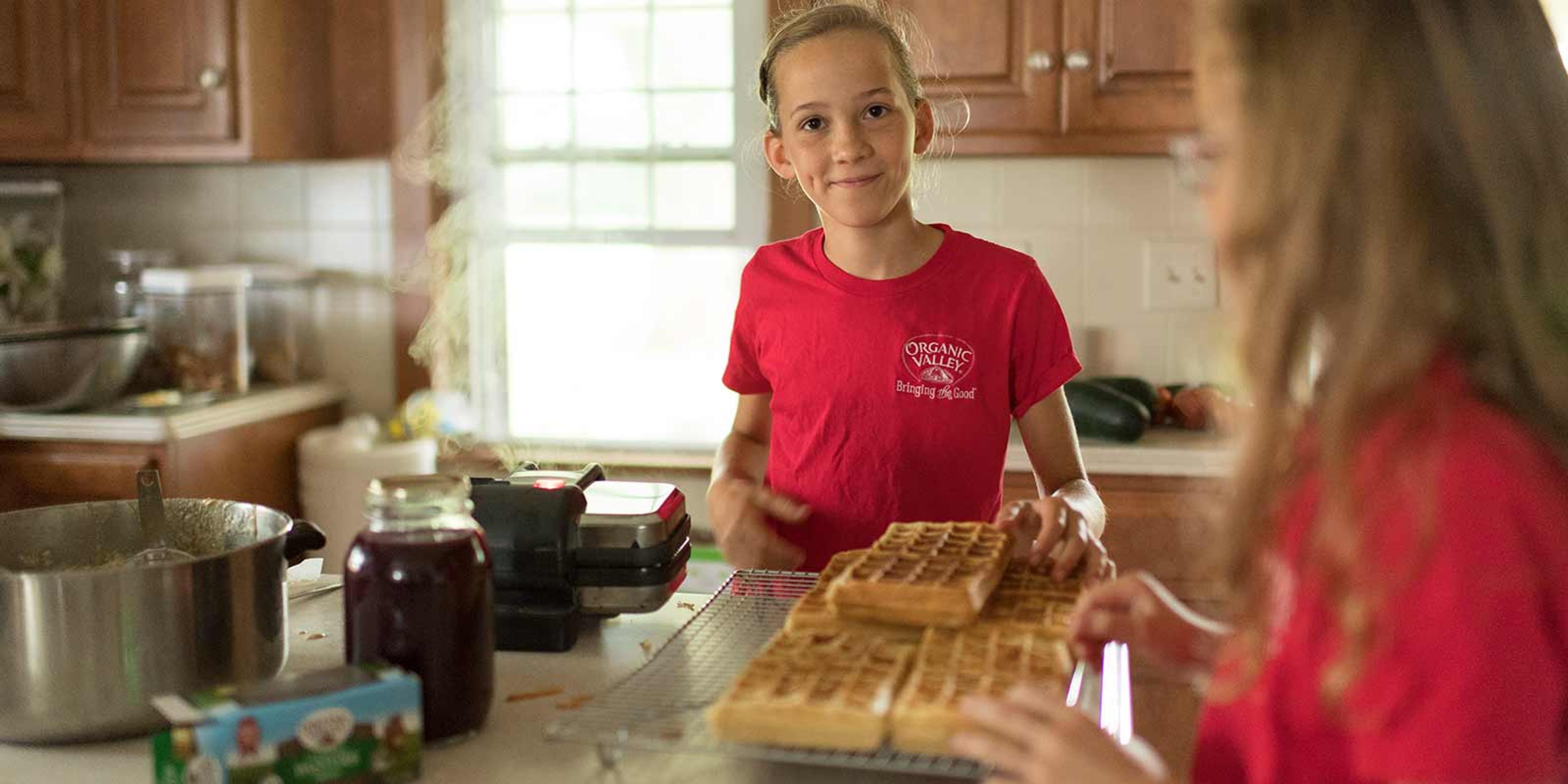 A girl wearing a red shirt smiles at the camera while standing in front of a pile of freshly made waffles in her family’s kitchen