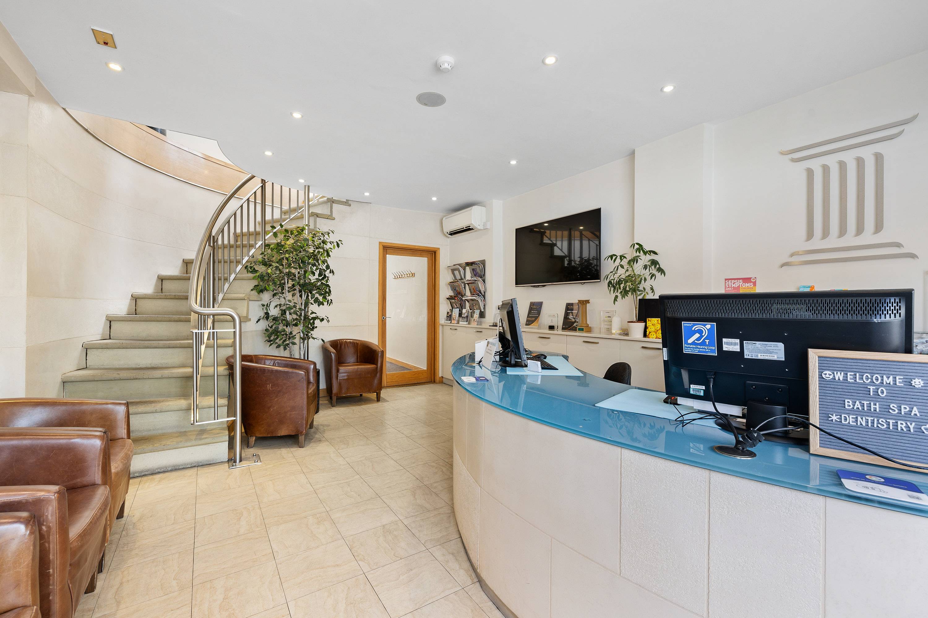 Reception and waiting area at Bath Spa Dentistry featuring a curved welcome desk, comfortable seating, and a staircase leading to treatment rooms.