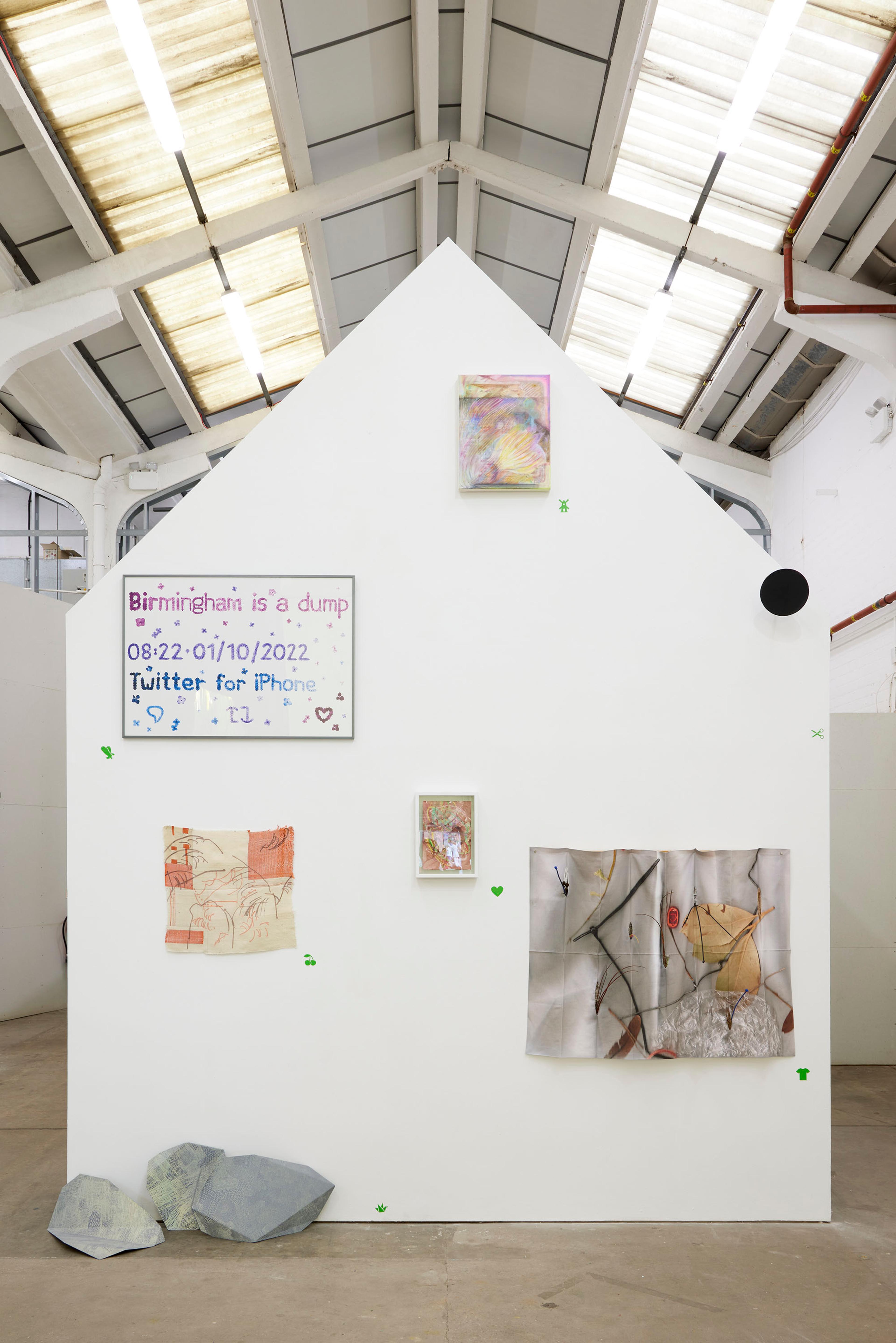 A house like structure in Eastside Projects, inside a warehouse space, a building within a building. The gable end is painted white and is the display structure for seven artworks by EOP members. On the floor on the bottom left leaning against the house wall are three drawings/sculptures. Grey rock like forms made of painted aluminium and covered all over with an intricate drawing in retroreflective pigment. The photographer has used his flash so they are gently lit up.  