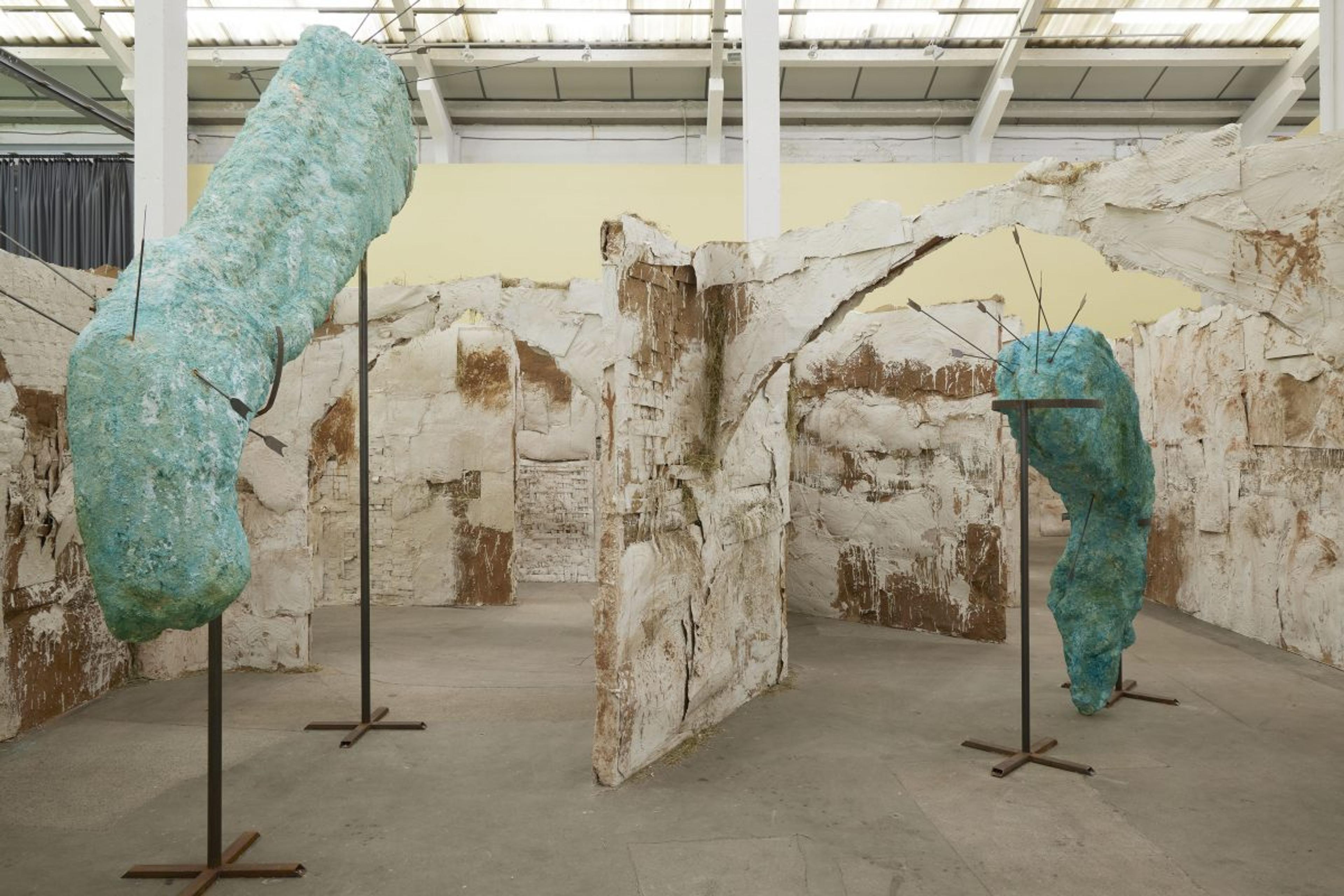 A view of Emii Alrai's large scale installation the Courtship of Giants. Two large turquoise sculptures coloured with copper oxide sit on large stands like large historical artefacts in museums. They are punctured with steel arrows. They sit inside a large maze made of layers of plaster, clay, straw and cardboard interspersed with woven elements.