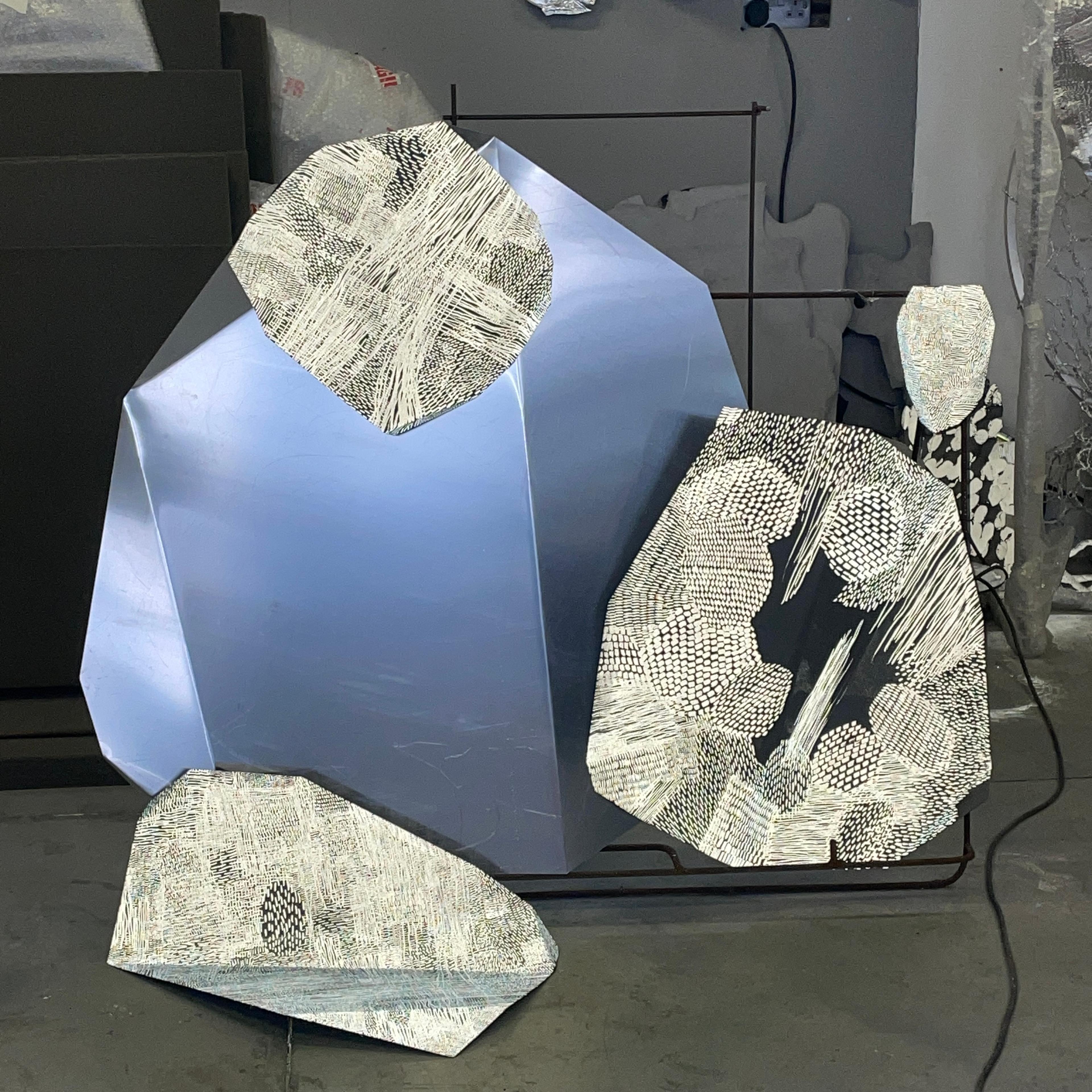 An arrangement of drawings on metal sheet lean against each other. At the back is a sheet of aluminium that is cut into a circular shape with the edges cut off to make it more angular and has three folds in it to give it facets. It's about a metre in diameter and has a blue colour on its surface which looks a bit like a blue sky. Hanging off the top, and leaning to the right and in the front on the left are three drawings on smaller pieces of painted aluminium cut into similar shapes and bent in several places. The drawings are made up of repetitive marks, like stitching across the surfaces. They are painted in retro-reflective pigment and then photographed with a flash on the camera so are glowing white.