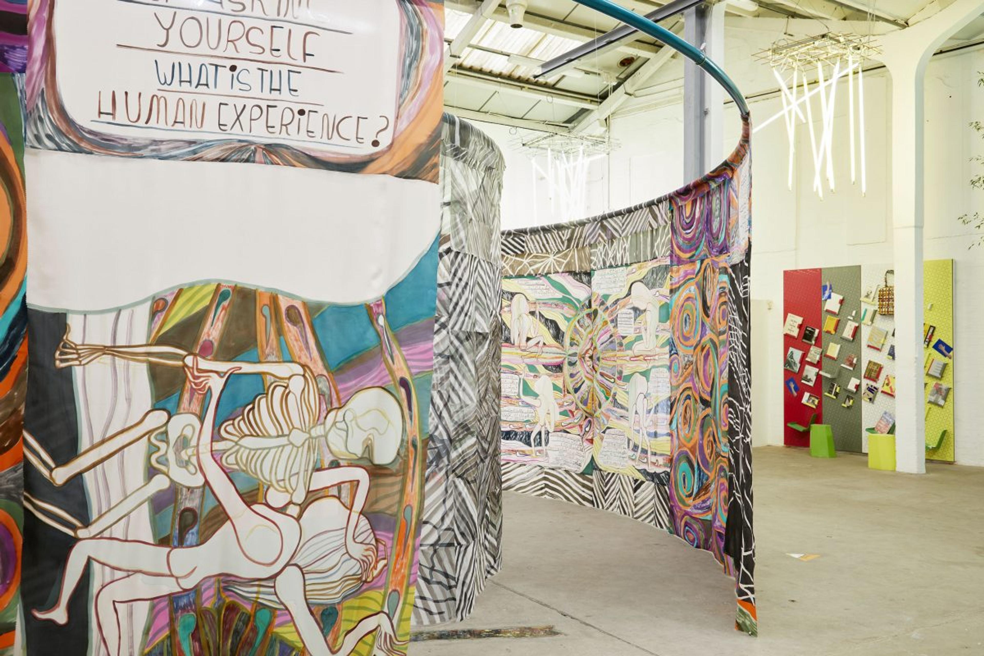 A large silk painting hands from a steel loop in the centre of Eastside Projects largest gallery. The paintings are brightly coloured and combine images, patterned panels and texts which ask questions like "What is the human experience". In the background you can see a bookcase by Martino Gamper and hanging fluorescent lights which are part of a functional artwork by Gavin Wade.