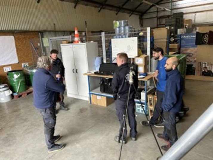 Six men in work clothes conversing in a warehouse.