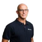 A bald man with glasses smiles slightly, wearing a dark blue polo shirt with a white logo on the chest.
