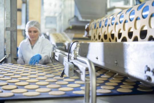 Opératrice en charlotte et blouse contrôlant la production de biscuits sur une ligne de fabrication agroalimentaire.