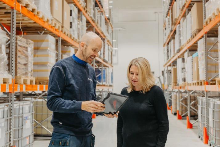 A man shows a tablet to a woman in a warehouse.