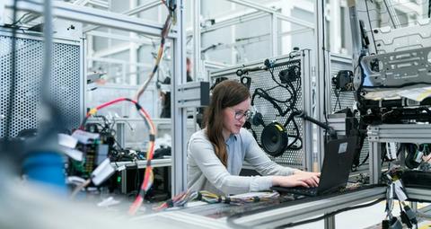 A female engineer in glasses types on a laptop amidst wiring harnesses and machinery in a modern factory.