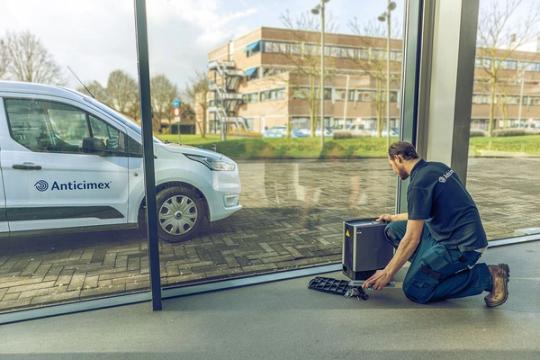 Anticimex technician kneeling to set up a device by a window, with a branded van outside.