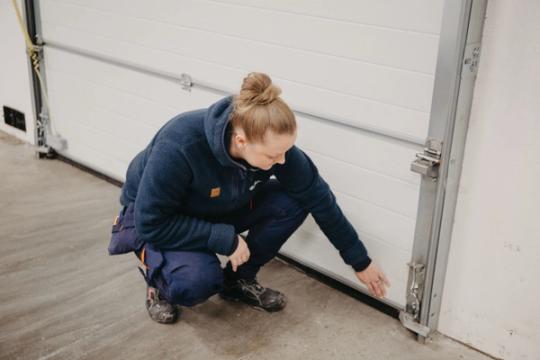 A woman in work clothes squats to inspect the bottom of a white garage door.