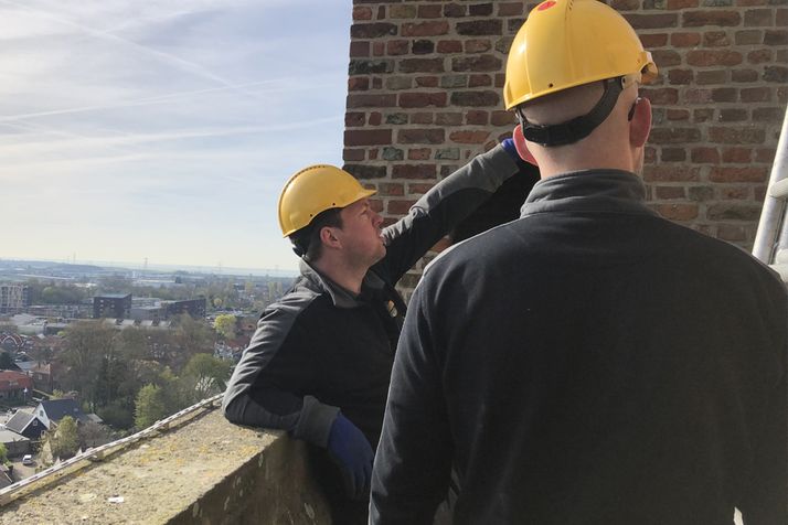Two men in yellow hard hats inspect a brick wall from a height, overlooking a city.