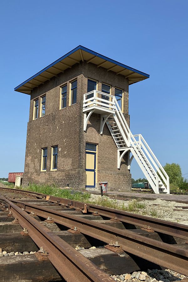 A three-story brick signal box with a blue-trimmed roof and white exterior stairs stands beside rusty train tracks under a clear blue sky.