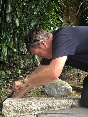 A man kneels, using a tool to work on a stone border outdoors.