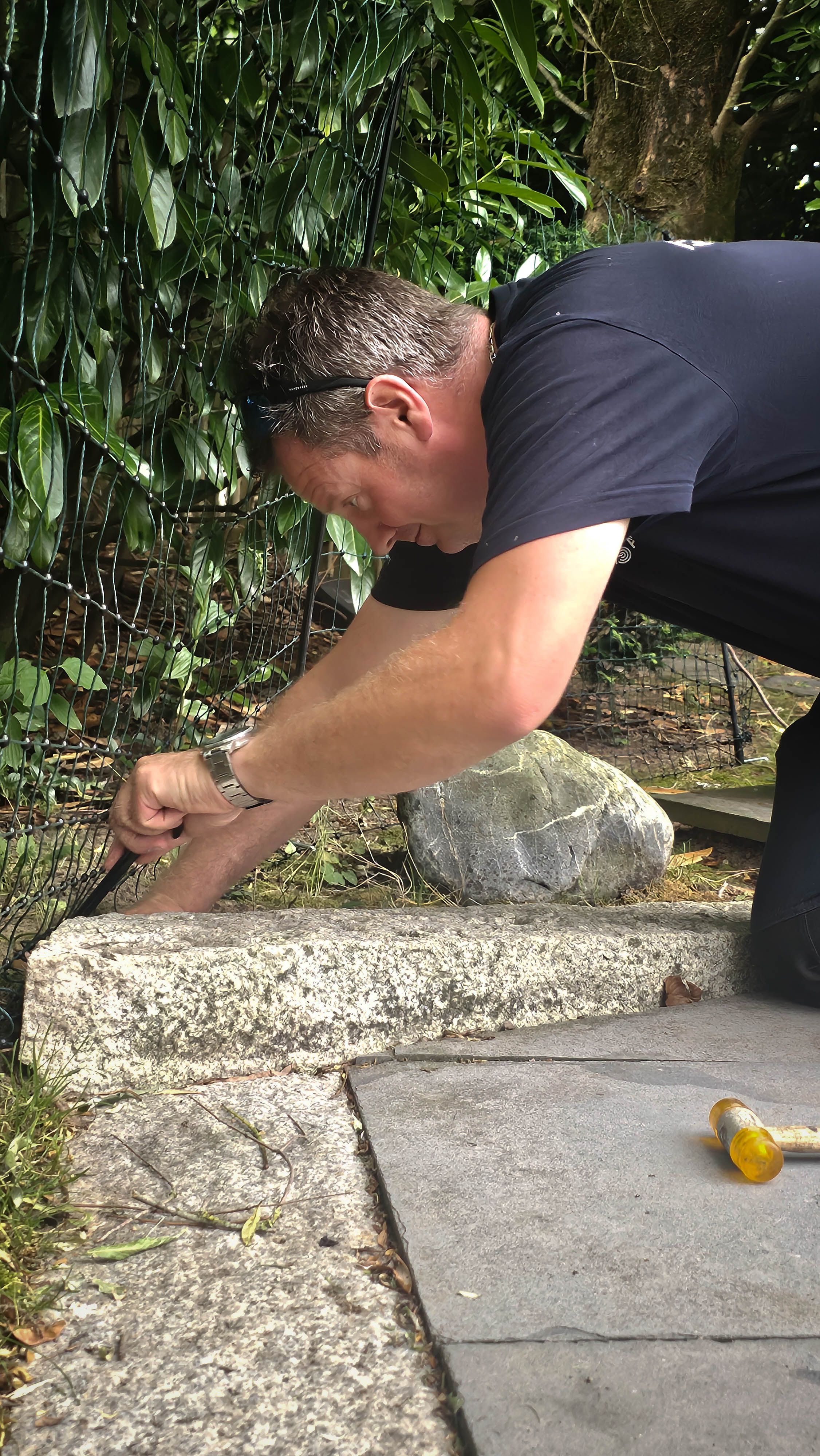 A man kneels, using a tool to work on a stone border outdoors.