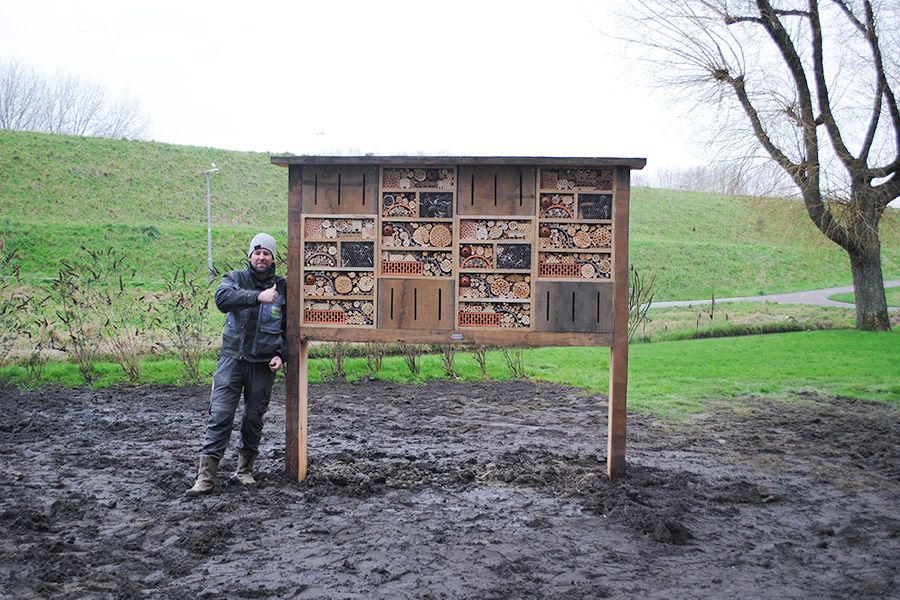 A man gives a thumbs-up next to a large insect hotel on muddy ground.