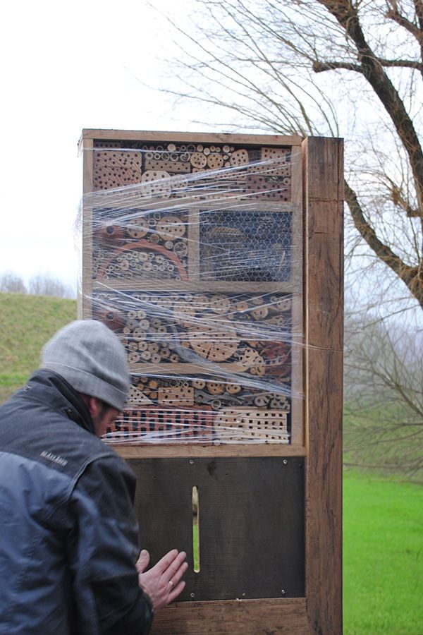 A person faces a large wooden insect hotel, wrapped in plastic and filled with various natural materials, in an outdoor setting.