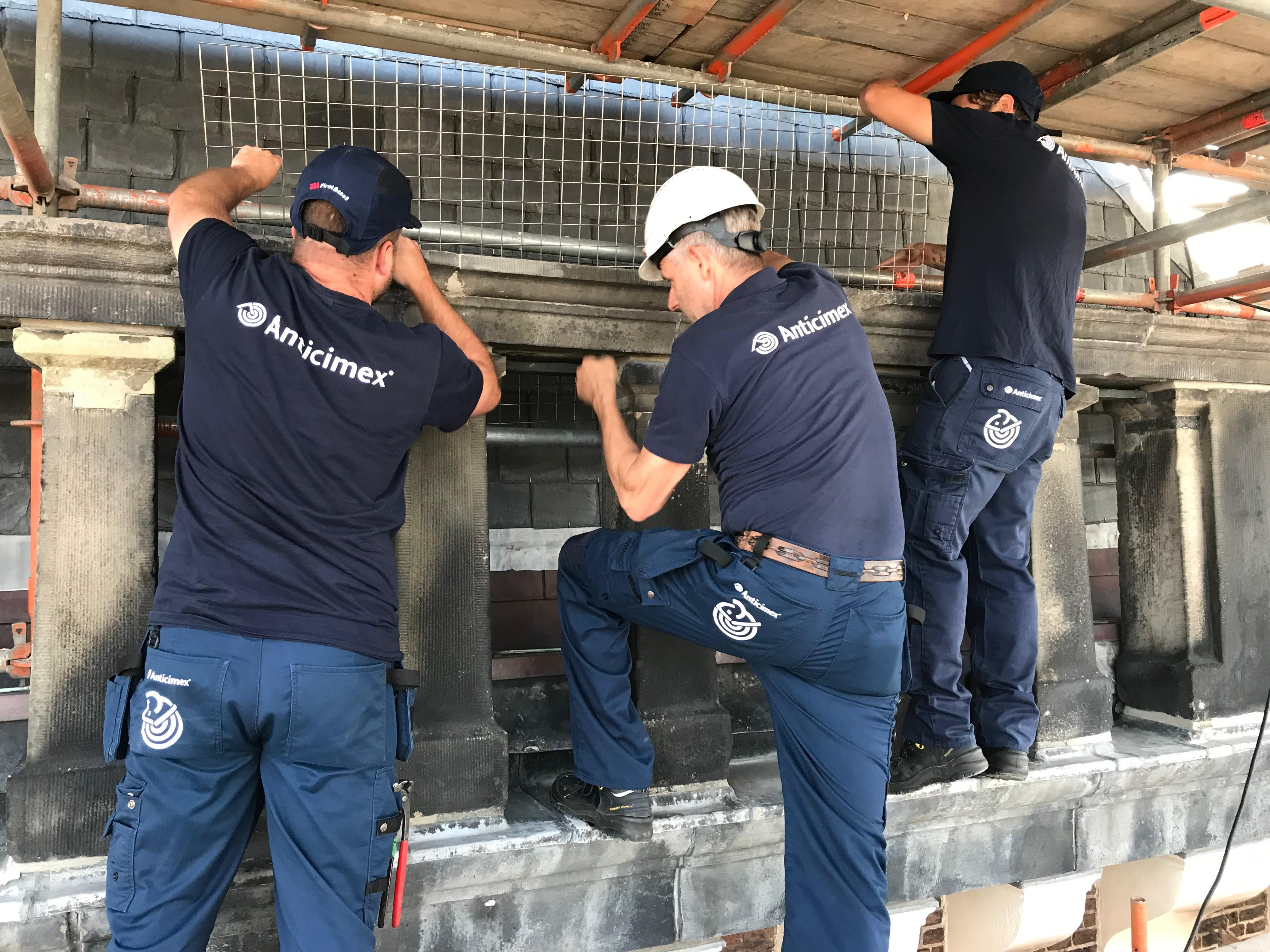 Three workers in Amicimex uniforms install wire mesh on a stone building from scaffolding.