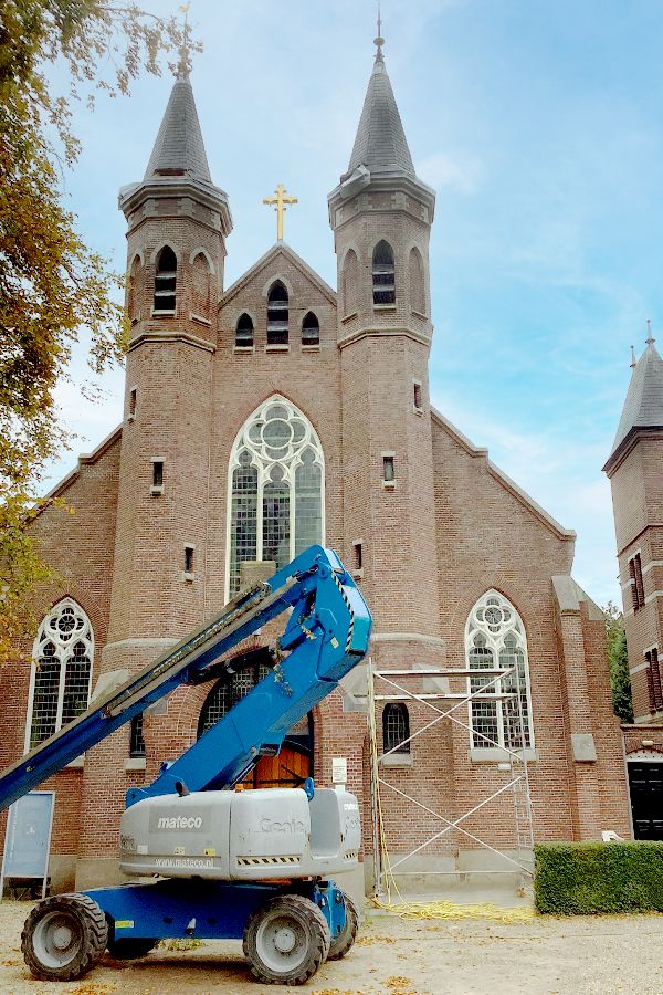 A brick church with two spired towers and a golden cross, with a blue boom lift and scaffolding in the foreground.