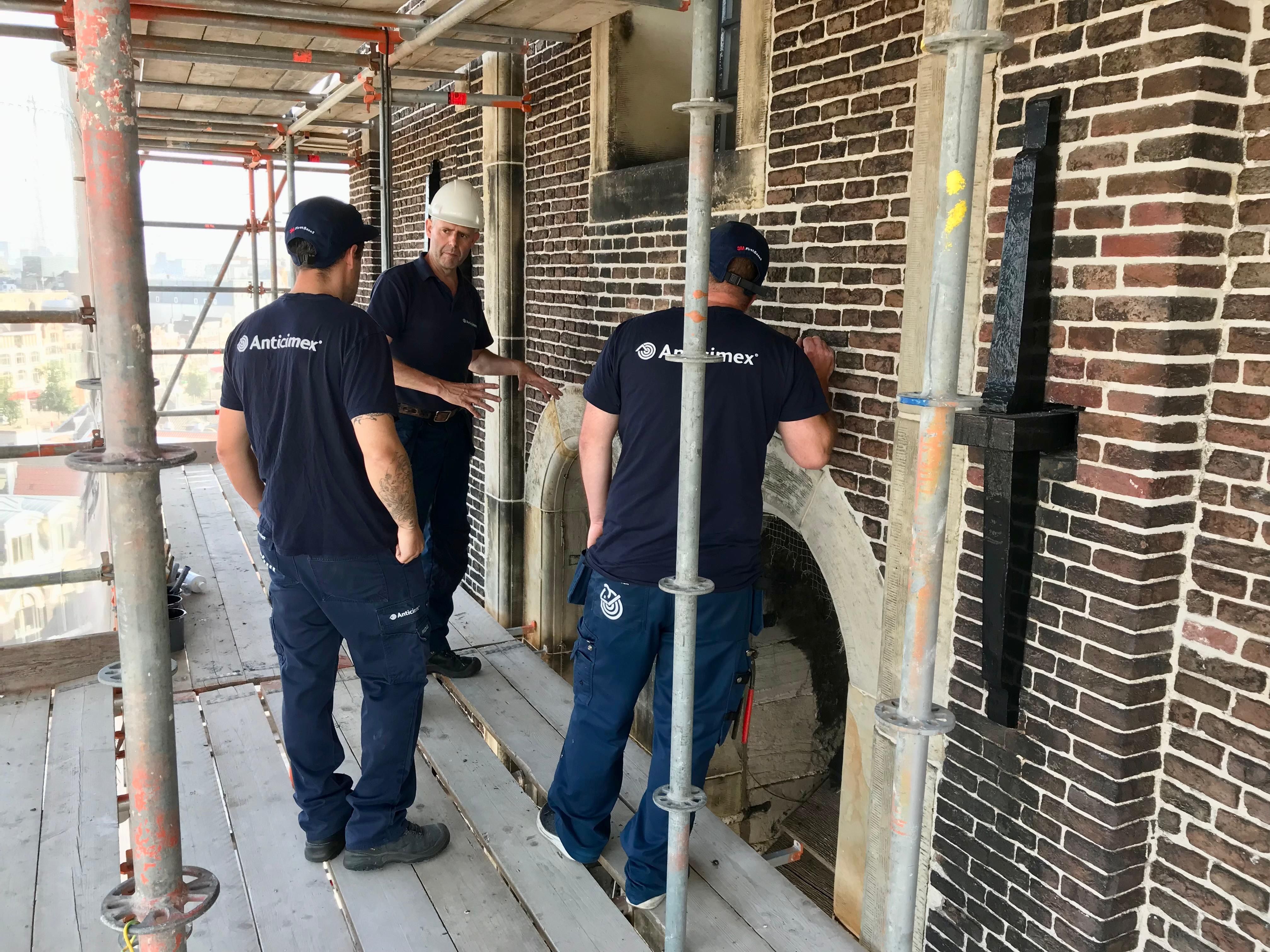 Three construction workers on scaffolding examine a brick building with an arched opening.