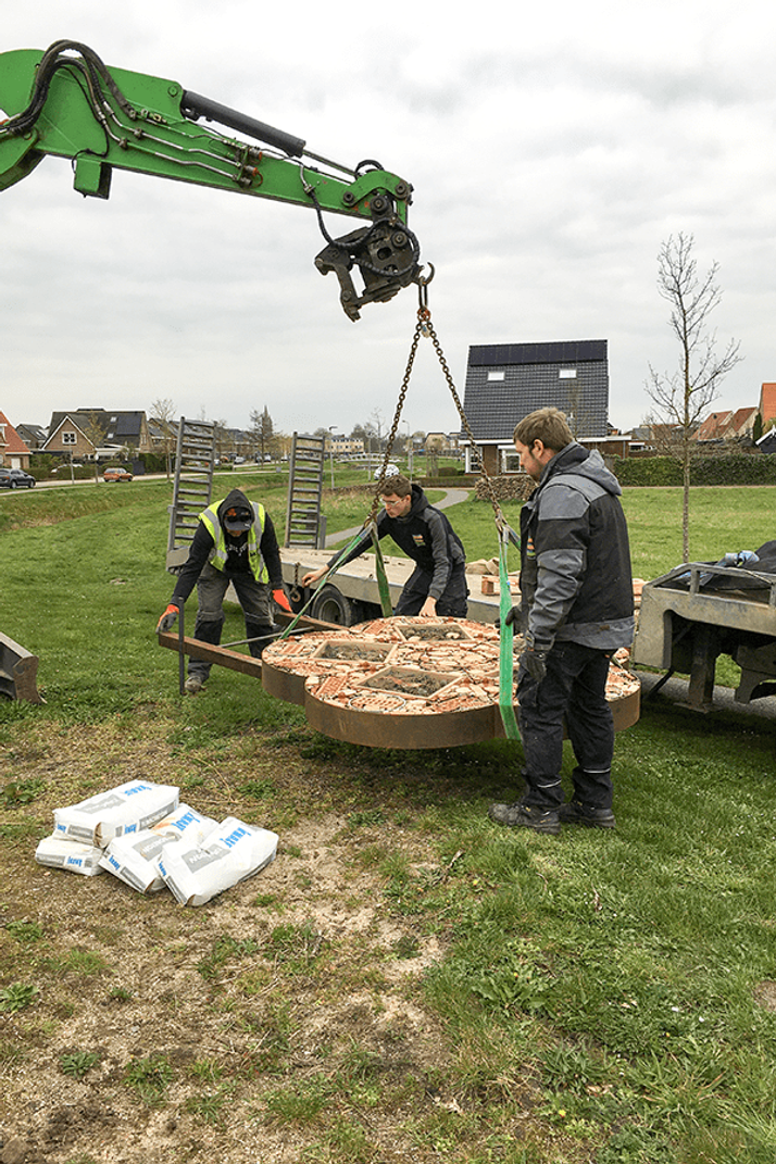 A crane lifts a large, circular brick-patterned disc, guided by three workers in a grassy area.