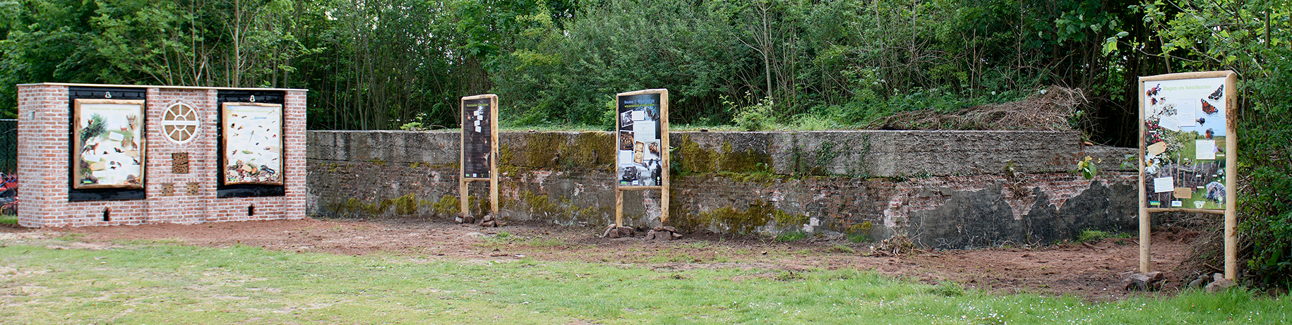 An outdoor interpretive display with information panels on a brick wall and freestanding signs, set against a low concrete wall and trees.