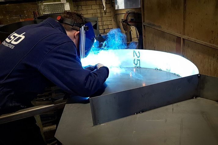 A person in a welding helmet and blue uniform welding metal, emitting bright blue sparks.