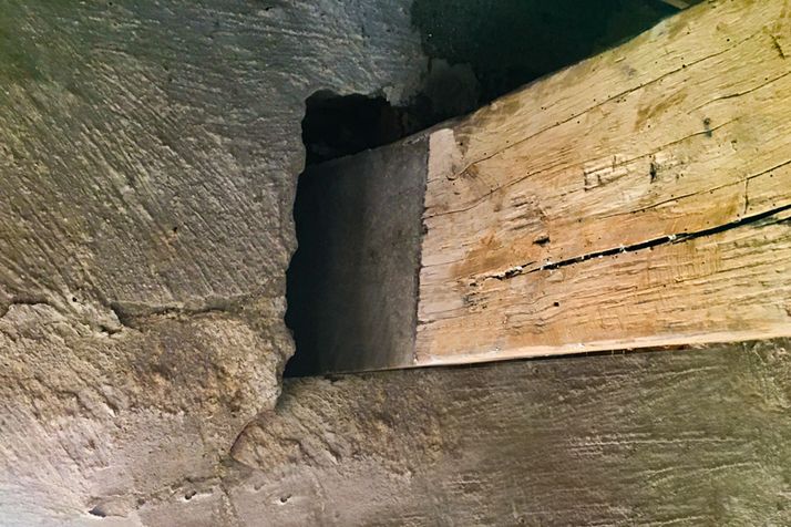 Rough stone wall with a dark opening, next to a weathered wooden beam with a long crack.