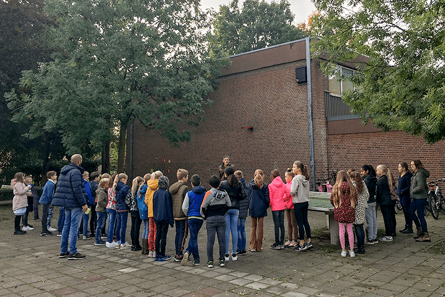 A group of students and adults stand on a paved area outside a brick building, listening to a person.