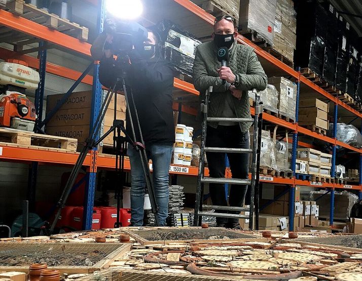 A reporter holding a microphone stands on a ladder next to a cameraman filming in a warehouse with tall shelves, over a grid of old, fragmented terracotta tiles and mesh.