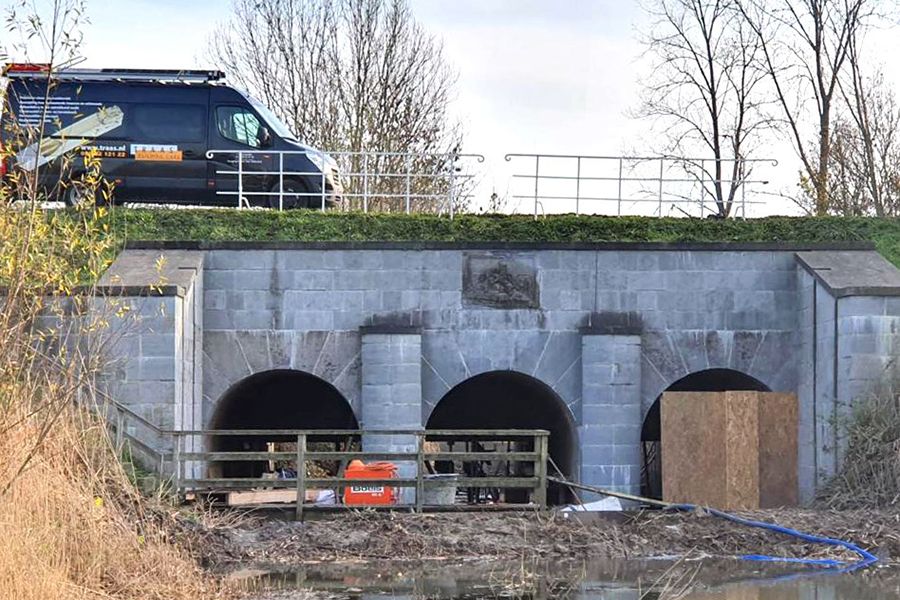 A three-arched stone bridge over a waterway, with a black van above and construction materials below.