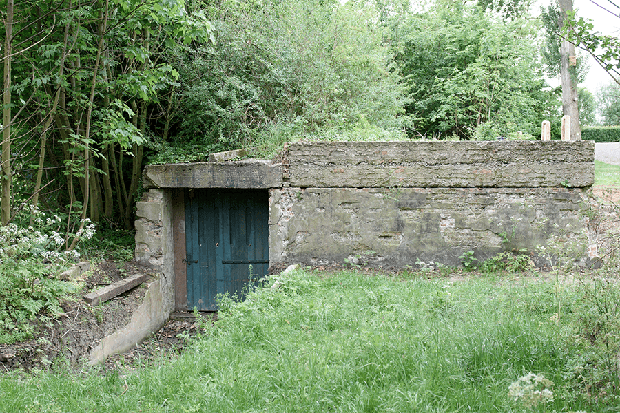 An old concrete structure with a green wooden door, set into an earthen bank and surrounded by trees and grass.