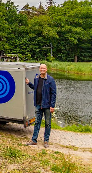 Three smiling men stand next to a black van and a grey trailer, both branded with a blue Borexmex logo, outdoors by a pond.