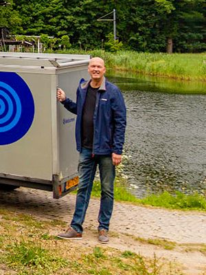 Three smiling men stand next to a black van and a grey trailer, both branded with a blue Borexmex logo, outdoors by a pond.
